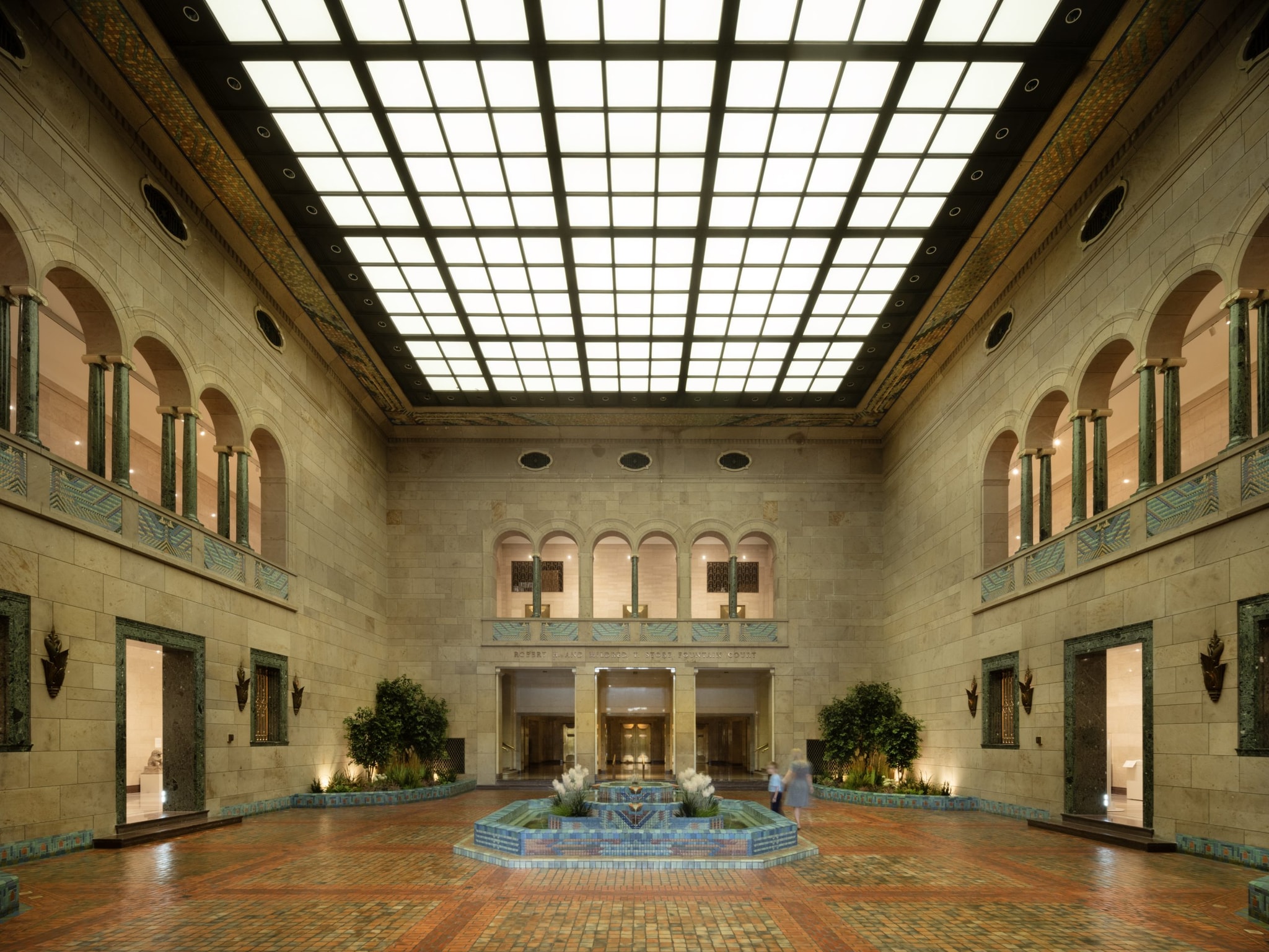 Interior of a large, ornate hall with a high glass-paneled ceiling. The room features archways, a symmetrical design, and a central fountain. The walls are adorned with decorative elements, and there are plants placed along the sides.