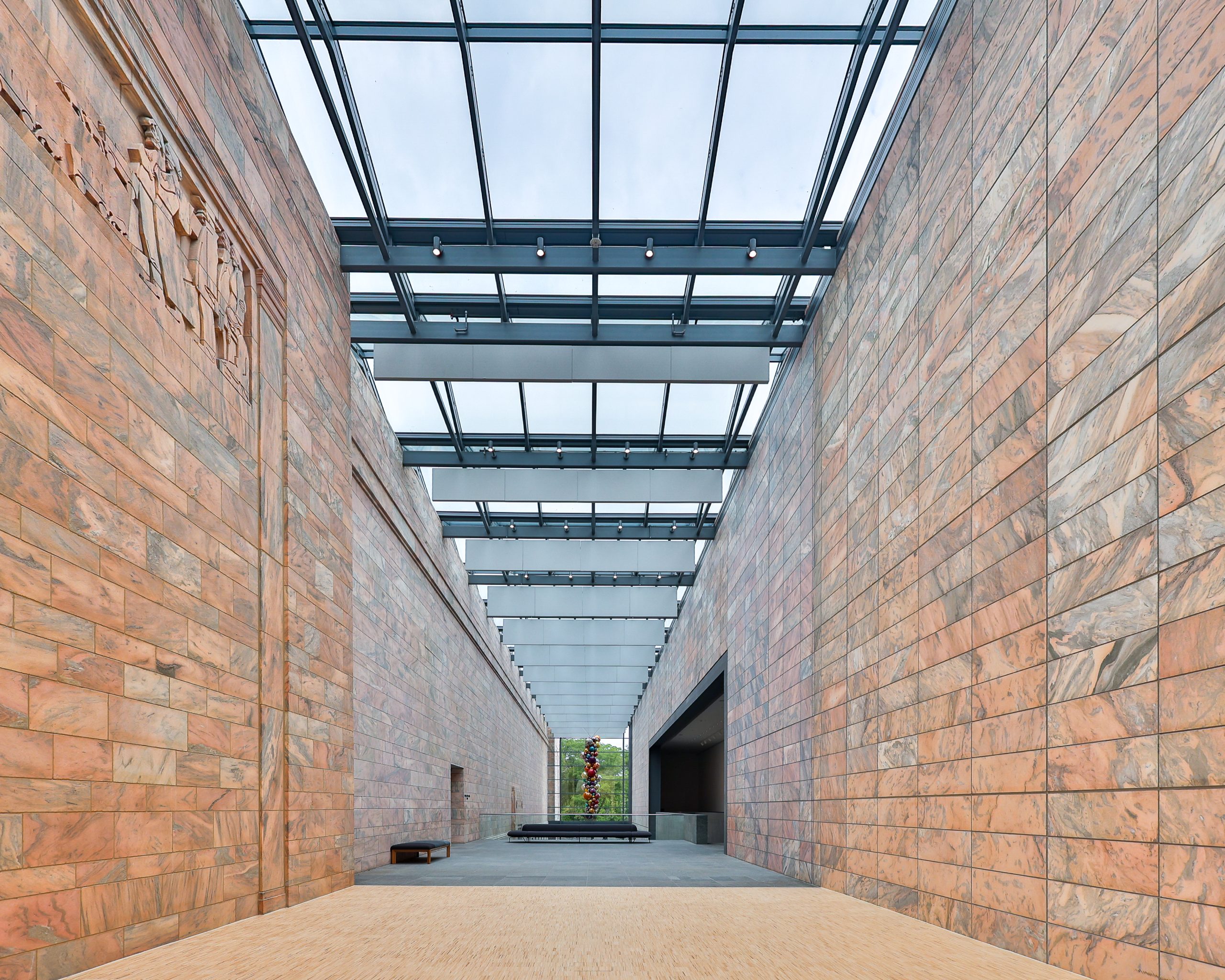 A modern architectural hallway with high glass ceilings flanked by tall stone walls. The passageway leads to a distant green courtyard, with a large sculpture visible in the center. Natural light illuminates the space.