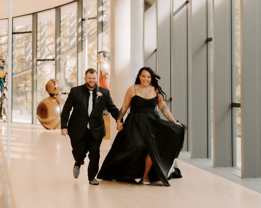 A couple is joyfully walking hand in hand down a sunlit glass corridor. The woman wears a flowing black dress and the man is in a black suit with a tie. Both are smiling as they move towards the camera.