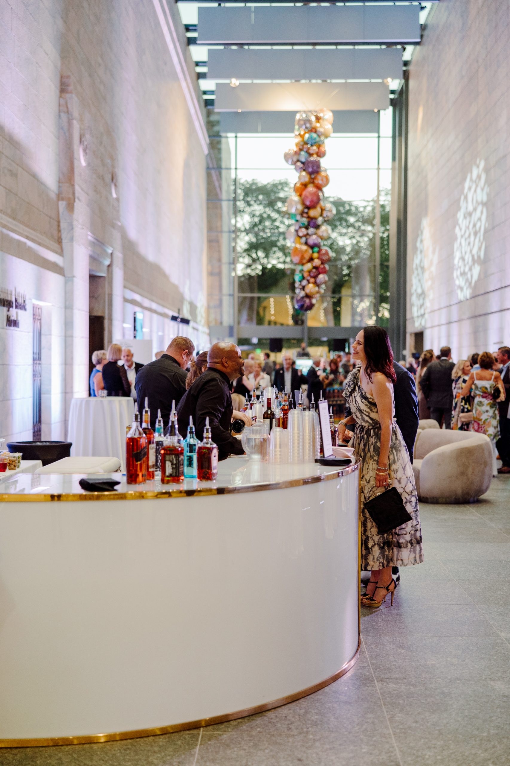 A stylish indoor event with a modern bar in the foreground, stocked with a variety of colorful drink bottles. People are mingling and talking in the background. A striking ceiling installation of suspended colorful spheres is visible above.