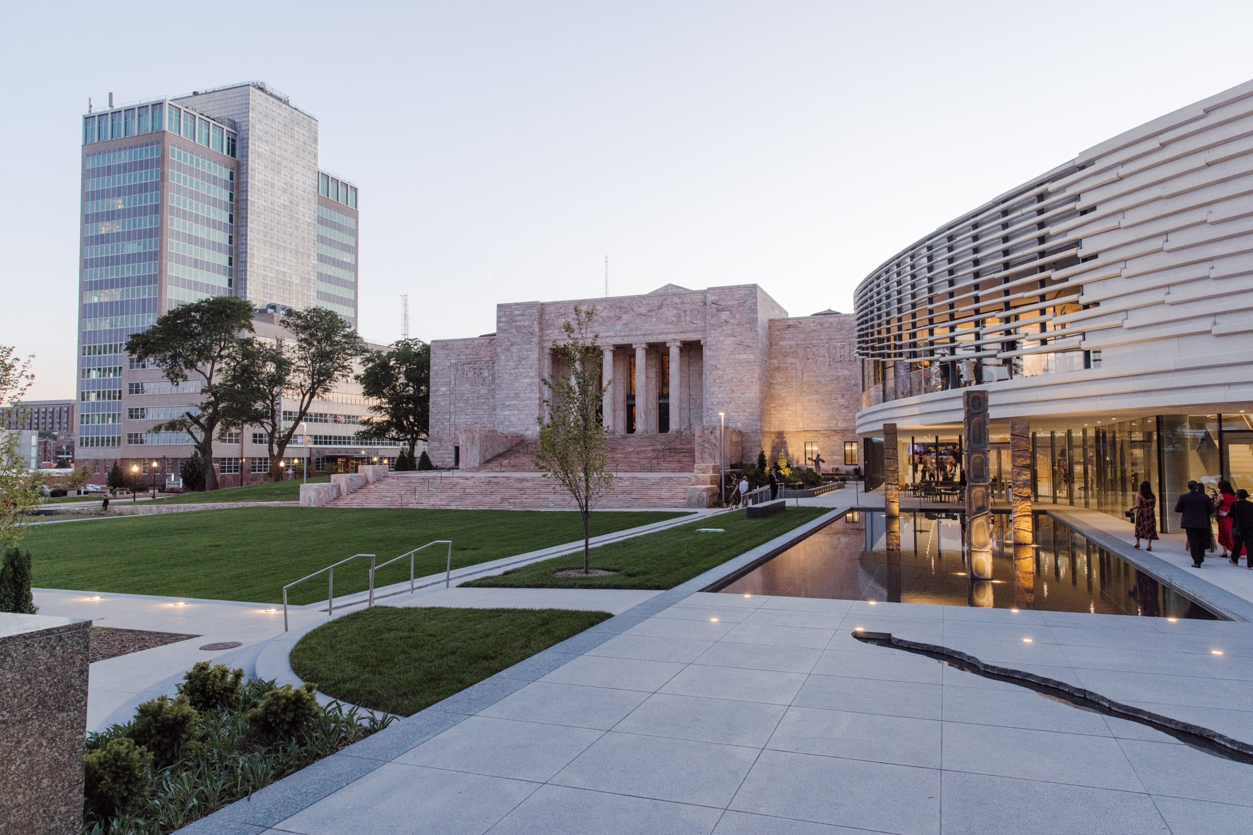 Outdoor plaza featuring a modern building with a curved facade and reflective windows next to a classic stone building with columns. A tranquil reflecting pool runs alongside. A tall office tower is in the background under a clear sky.