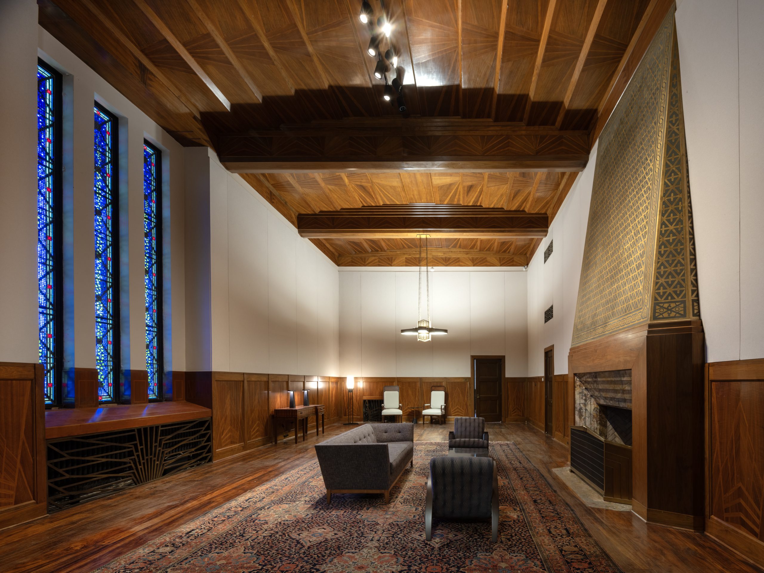 Spacious room with high wooden ceiling, featuring a geometric pattern. Stained glass windows on the left, large patterned rug on the floor, and modern furniture including gray couches and white chairs. A decorative fireplace is on the right wall.