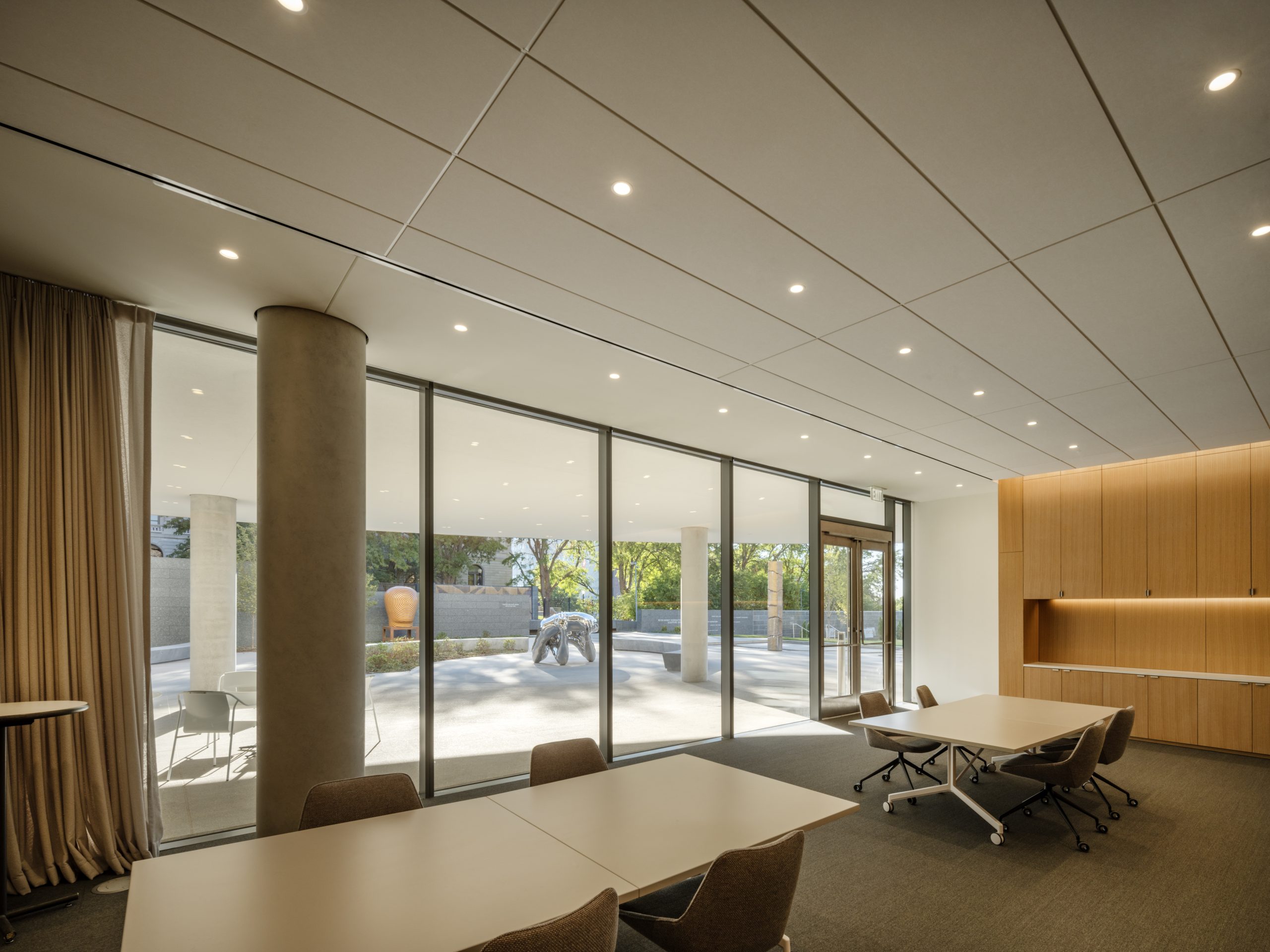 Modern conference room with large floor-to-ceiling windows offering a view of an outdoor area with trees. The room has tables, wheeled chairs, a wall unit, and beige curtains. Ceiling lights are evenly spaced.