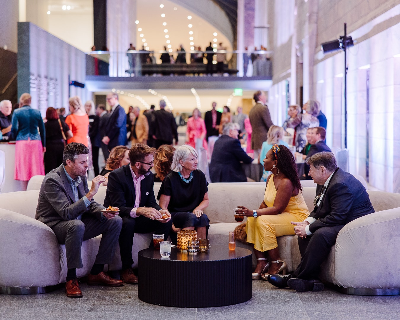 A group of five people sit on a curved sofa at a formal event, engaged in conversation. They are surrounded by other guests mingling in a spacious, elegantly lit venue. Decorative candles and drinks are on the small table in front of them.