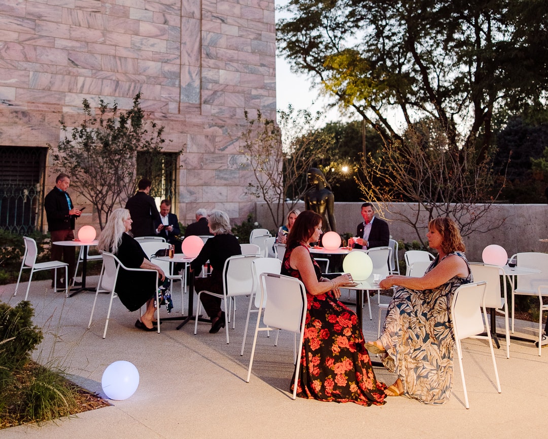 Outdoor evening gathering with people seated at tables adorned with glowing spheres. Two women in floral dresses are in the foreground. A large stone building and trees are in the background, creating a relaxed atmosphere.