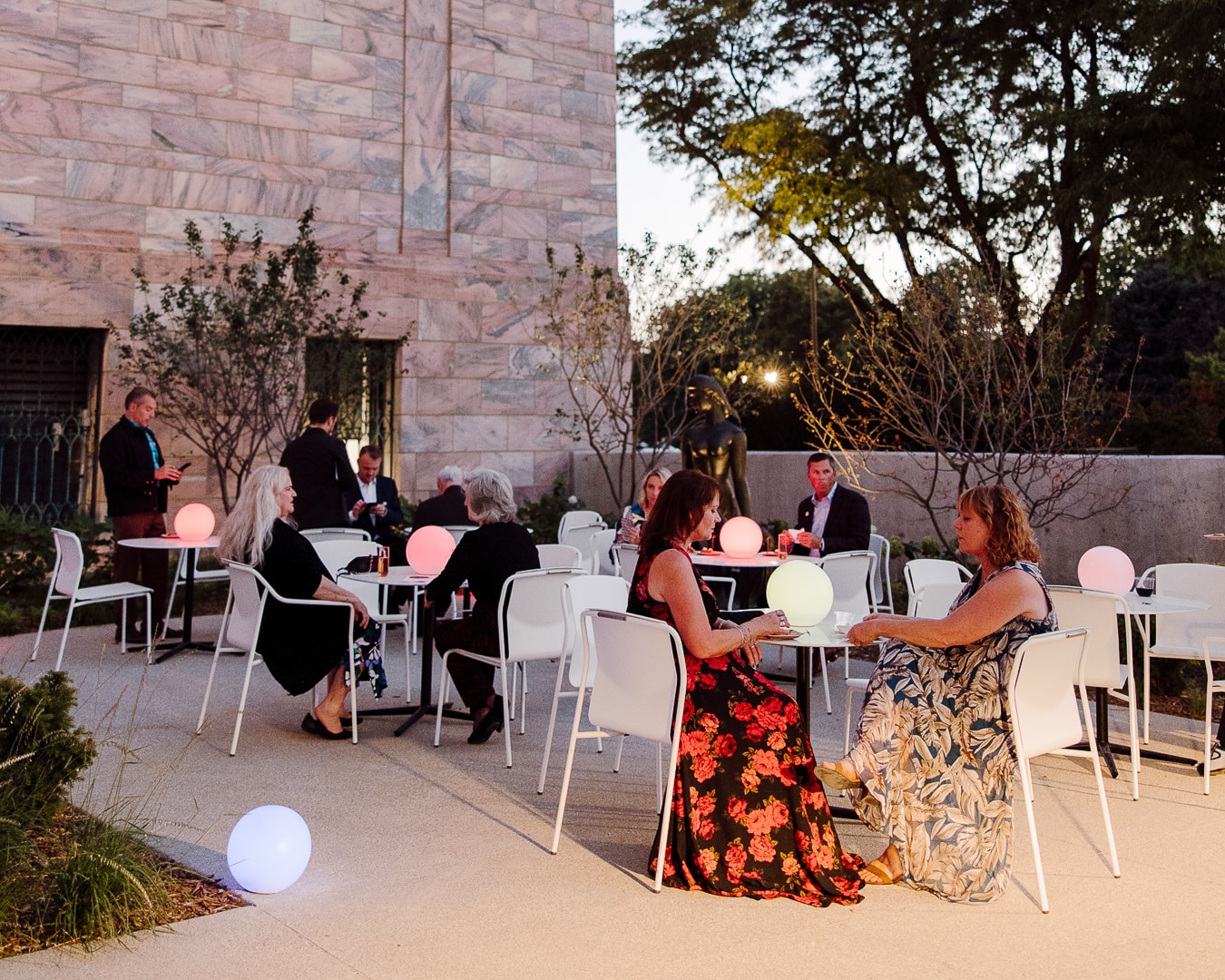 Outdoor evening gathering with people seated at tables adorned with glowing spheres. Two women in floral dresses are in the foreground. A large stone building and trees are in the background, creating a relaxed atmosphere.