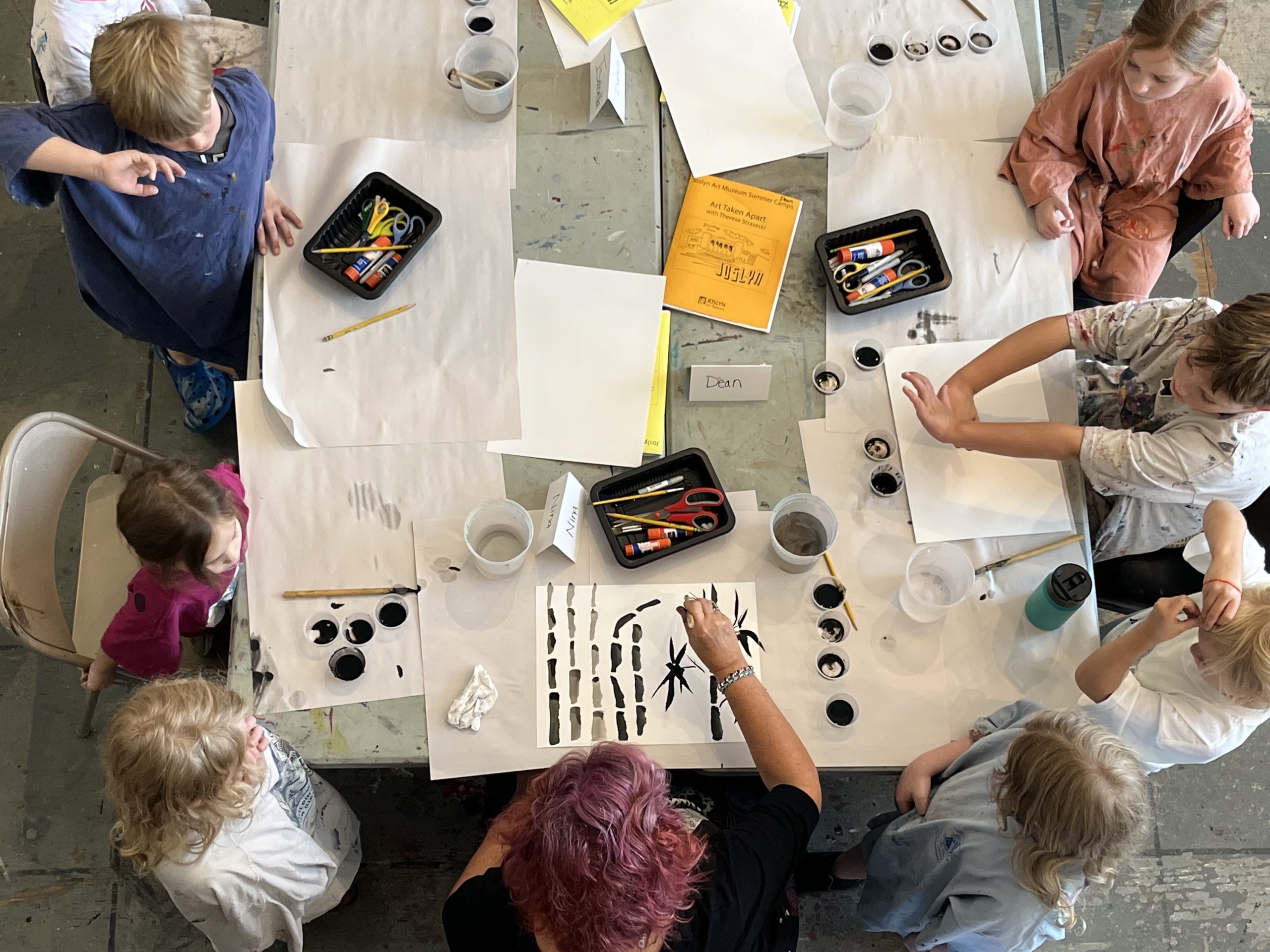 A group of children sit around a table as they are engaged in a painting activity. The table is covered with art supplies including brushes, containers of black paint, scissors, and papers with various designs. An adult supervises the activity.