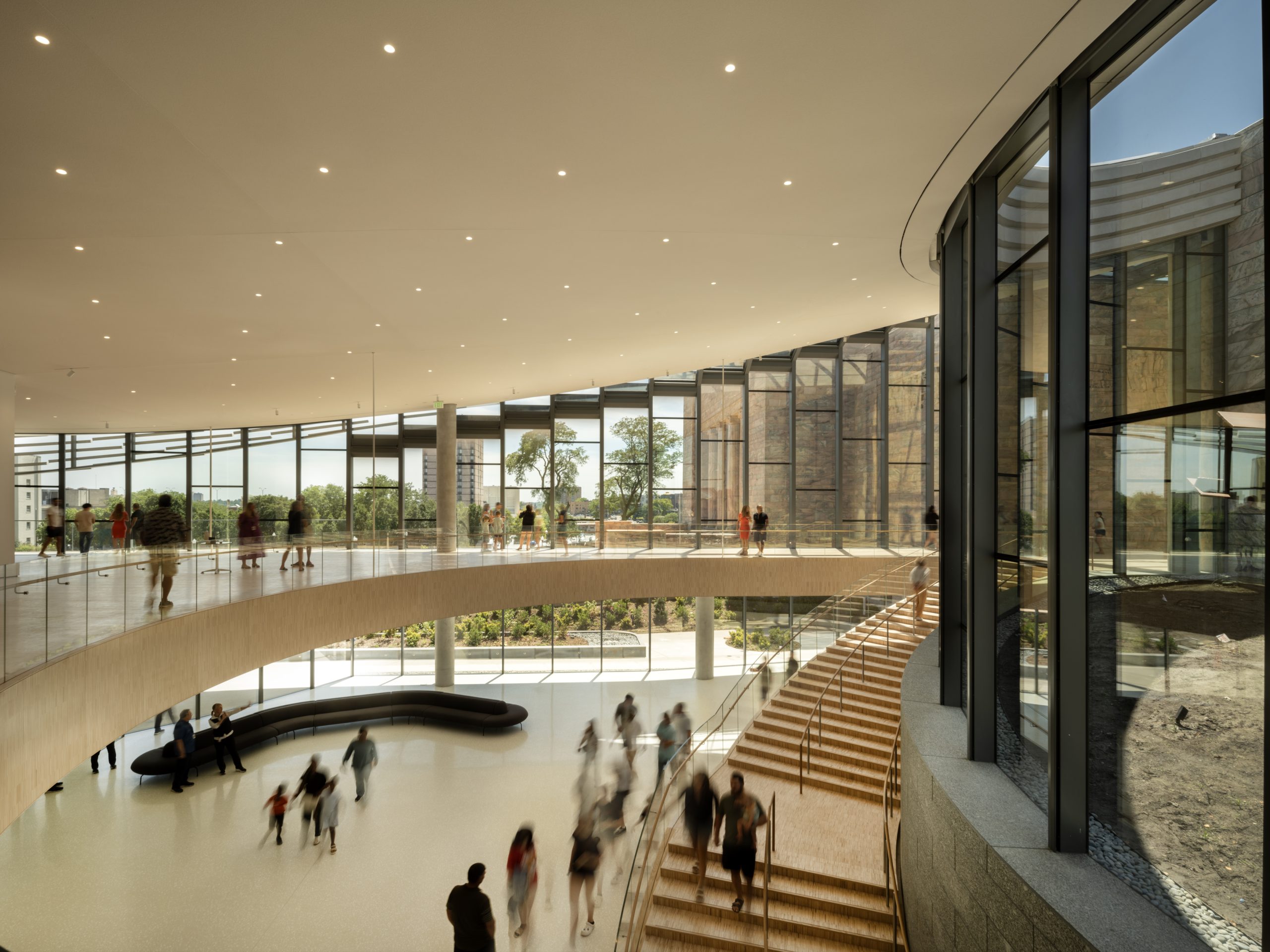 Spacious modern interior with large curved windows and a mezzanine. People walk on both levels, creating a sense of movement. Natural light streams in, highlighting the open, airy architectural design.