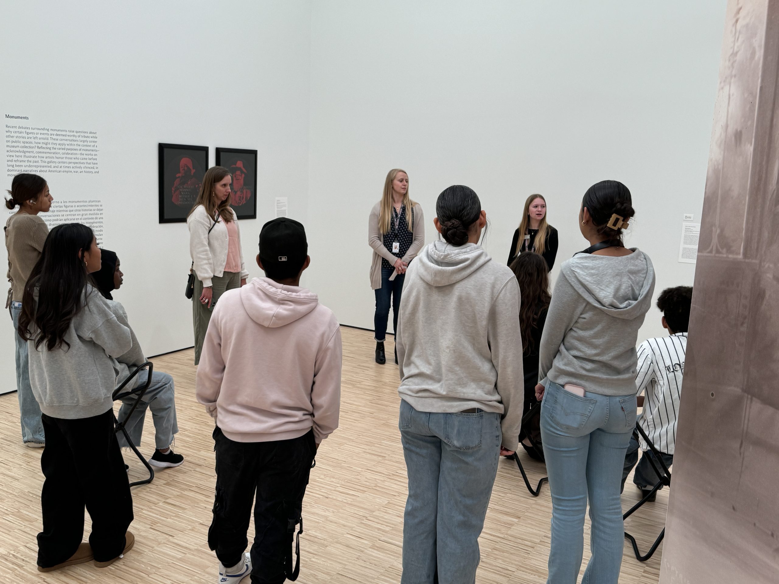 A group of people are gathered in an art gallery. Some are seated while others stand, facing two individuals presenting in front of two framed artworks on the wall. The room has light-colored wooden floors and white walls.