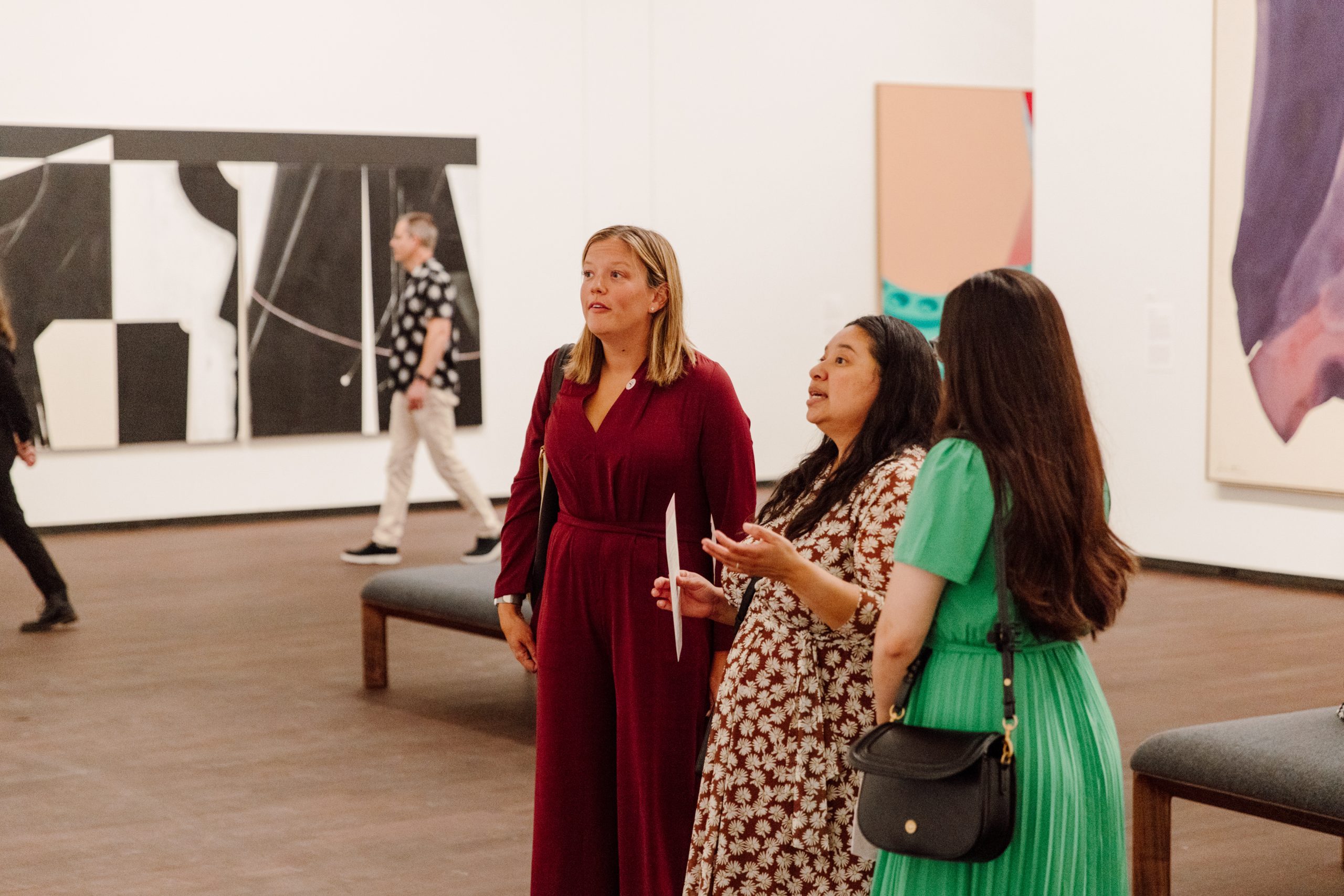 Three women stand in an art gallery, observing abstract paintings. One wears a red dress, the second a floral dress, and the third a green dress. In the background, a man walks past more artwork.
