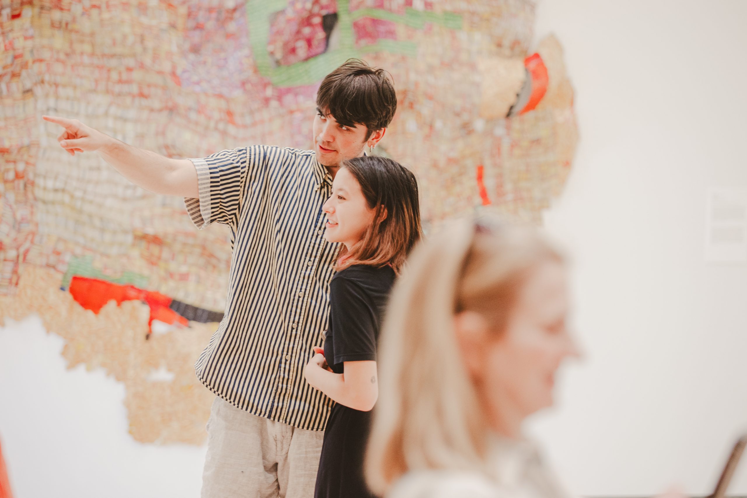 A young man and woman stand together in a gallery with the man pointing at artwork. They both appear engaged and smiling. A blurred figure is in the foreground, and an abstract wall piece is in the background.
