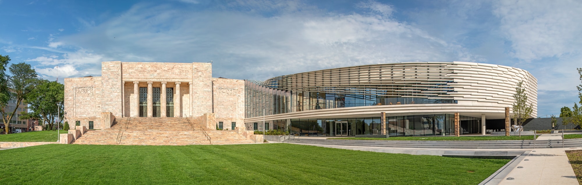 A panoramic view of a modern architectural building featuring both classical and contemporary elements. The structure includes stone steps leading to a façade with large windows and a circular section with horizontal slats, set against a blue sky.