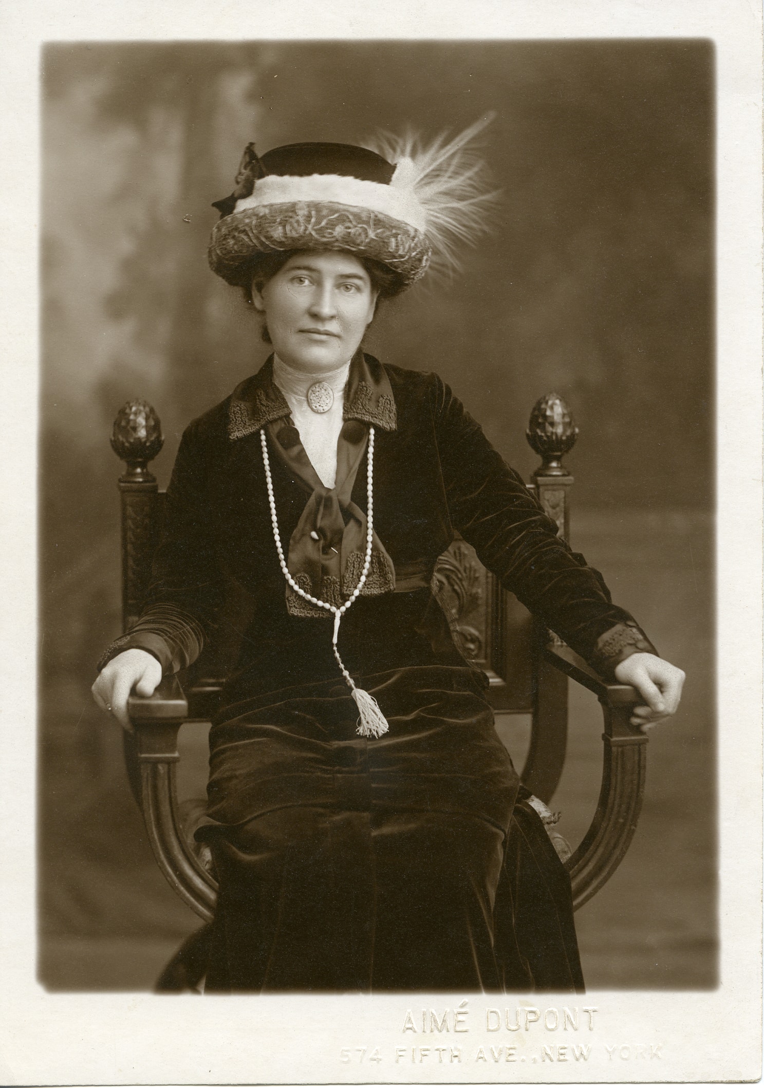 A vintage black and white portrait of a woman sitting on an ornate chair. She is wearing a dark dress, a pearl necklace, and a hat adorned with a feather. The bottom of the photograph reads "Aimé Dupont, 574 Fifth Ave., New York.