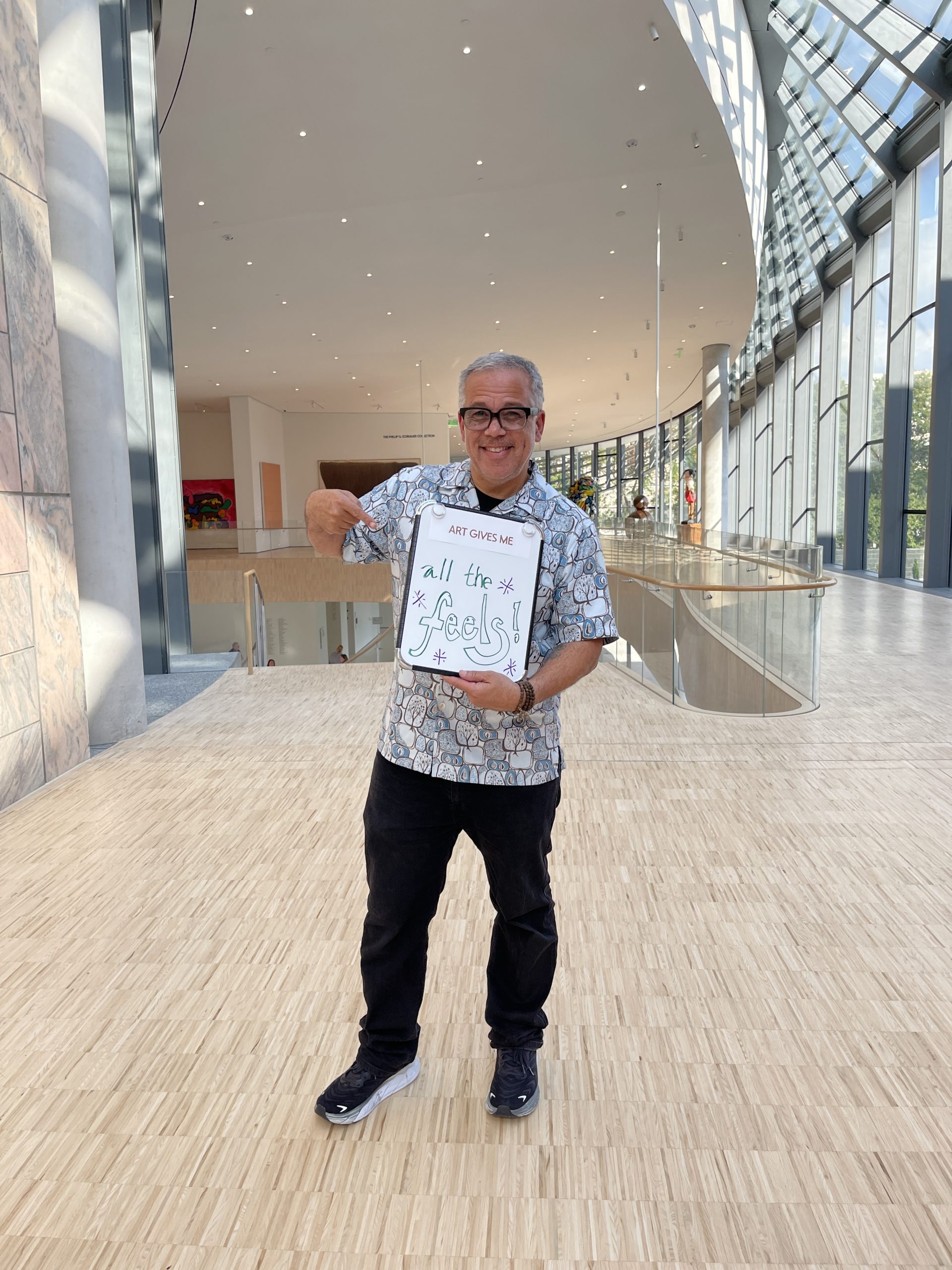 A person stands in a bright, modern building, smiling and holding a sign that says, "Art gives me all the feels!" The space features large windows and a wooden floor, creating an airy atmosphere.