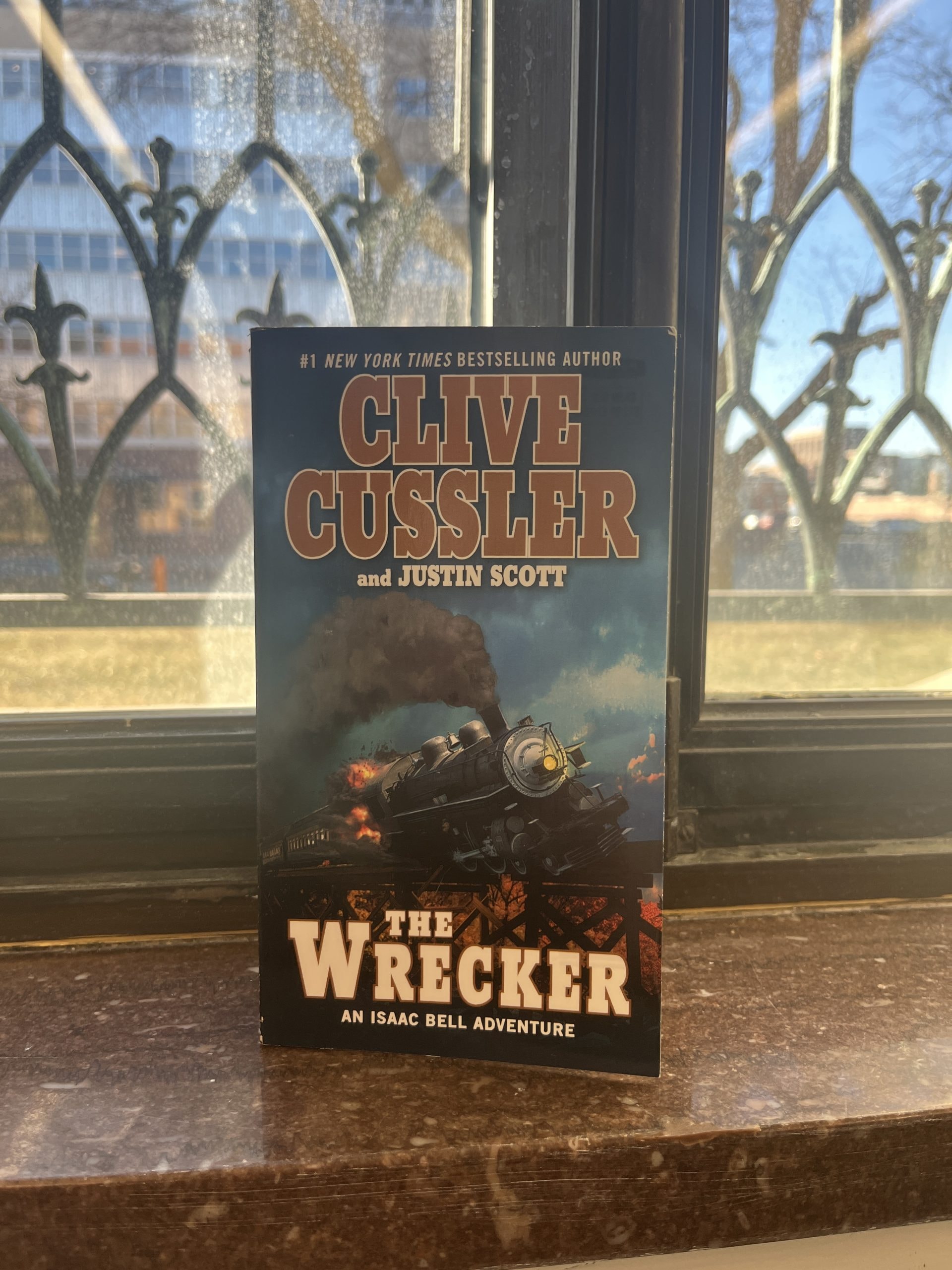A book titled "The Wrecker" by Clive Cussler and Justin Scott sits on a windowsill. The cover features a dramatic illustration of a train bursting through flames. A cityscape and iron window grilles are visible in the background.