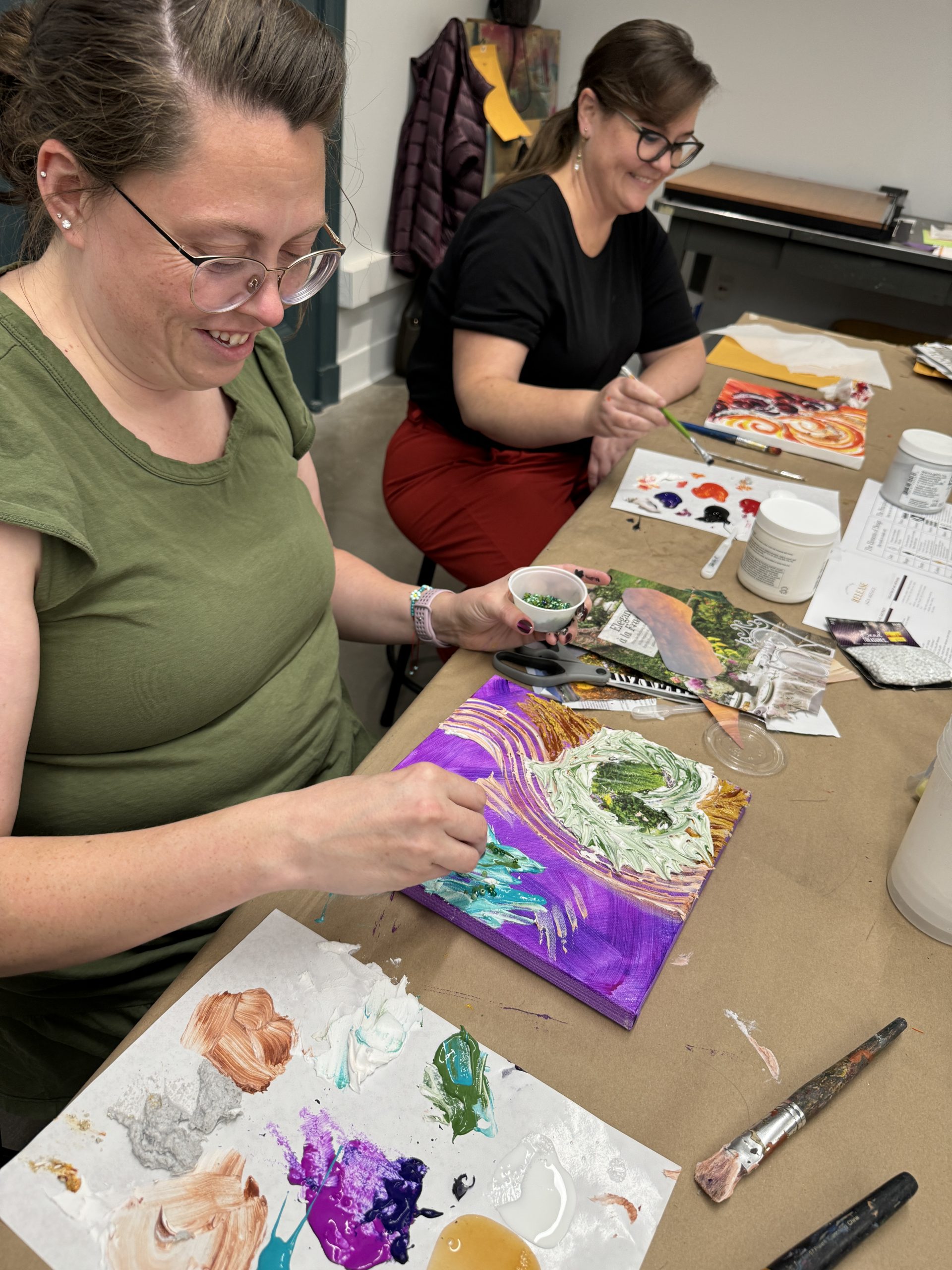 Two people are seated at a table, painting on small canvases. The person in front is smiling, wearing glasses and a green shirt. Various paint colors and brushes are on the table. The person in the back is also smiling, wearing glasses and a black shirt.