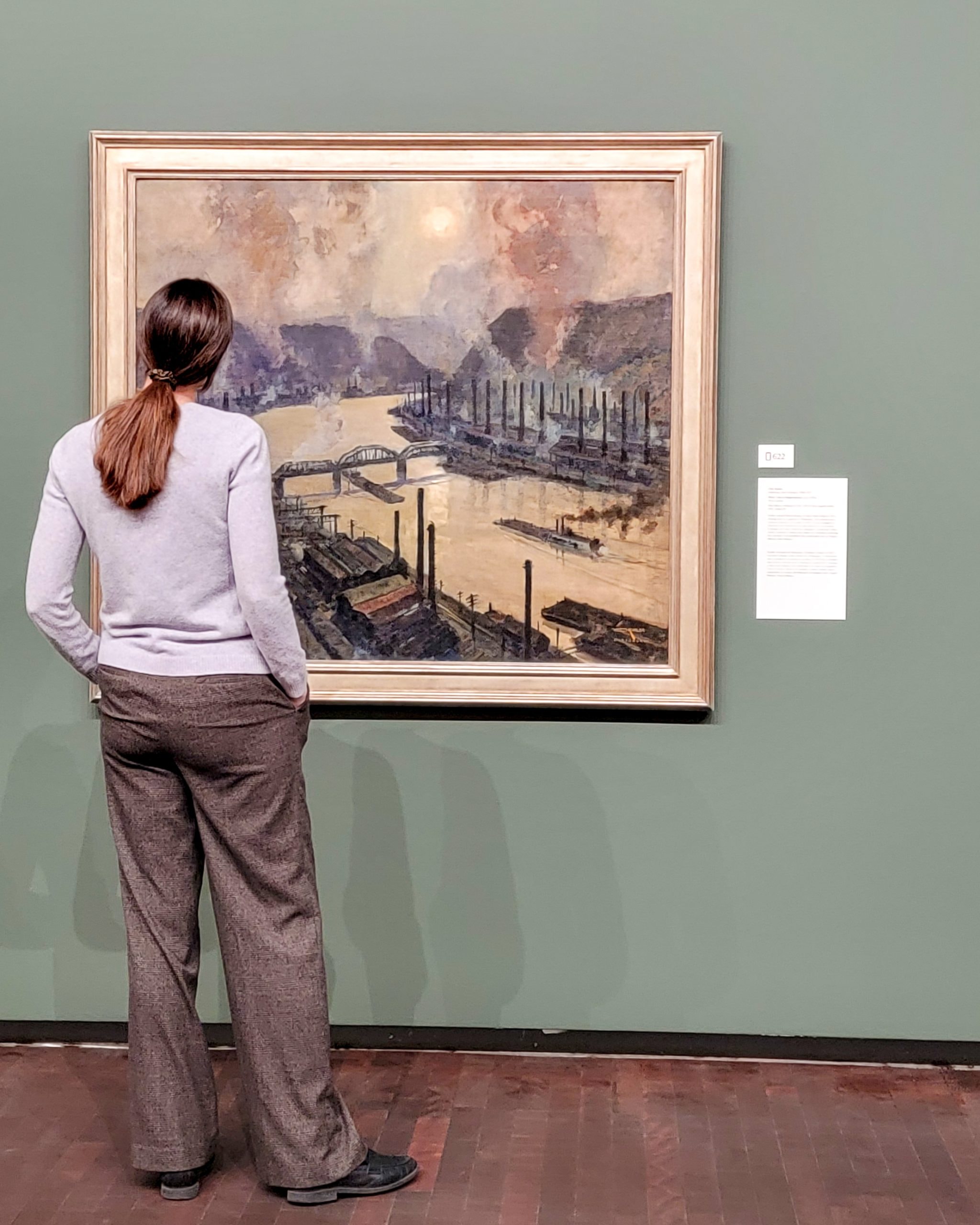 A person with long hair in a ponytail, wearing a gray sweater and brown pants, stands in front of a framed painting in a museum. The painting depicts a waterfront scene with boats and poles against a colorful sky.