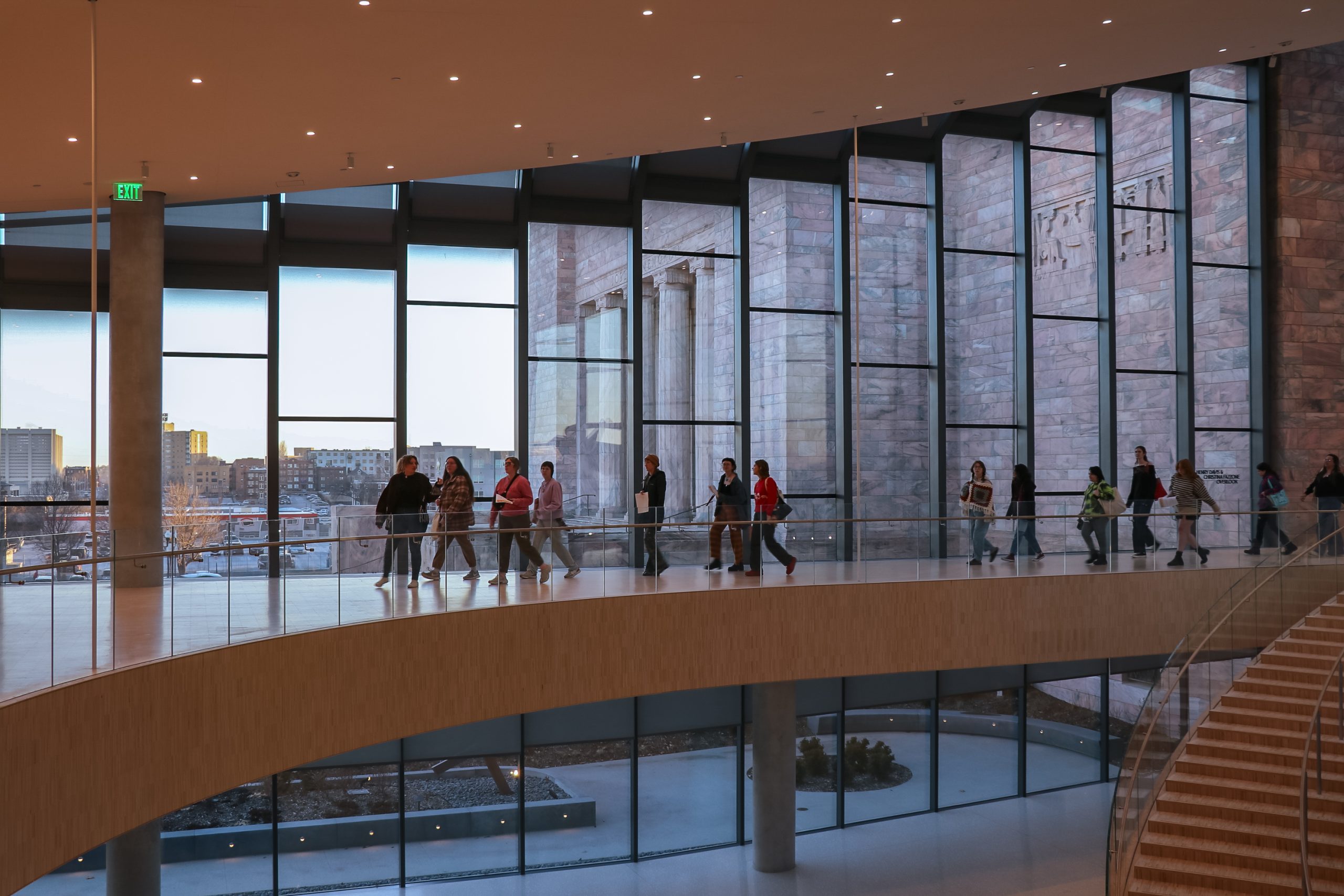 People walk along a curved glass walkway inside a modern building. The large floor-to-ceiling windows reveal a cityscape outside and a view of a stone wall. Warm interior lighting contrasts with the cool tones of the city view.