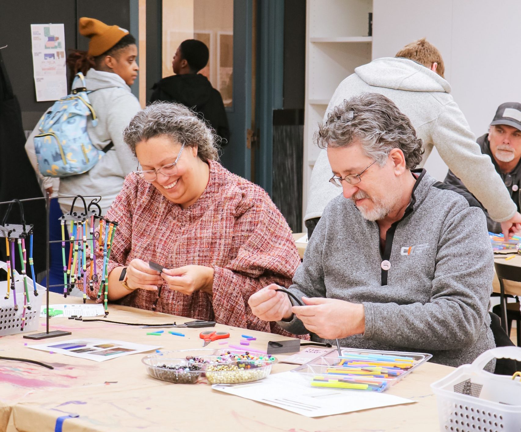 A group of people sitting at a table doing crafts. They are engaged with colorful materials like beads and markers. The room has craft supplies scattered on the table and people appear focused and cheerful.