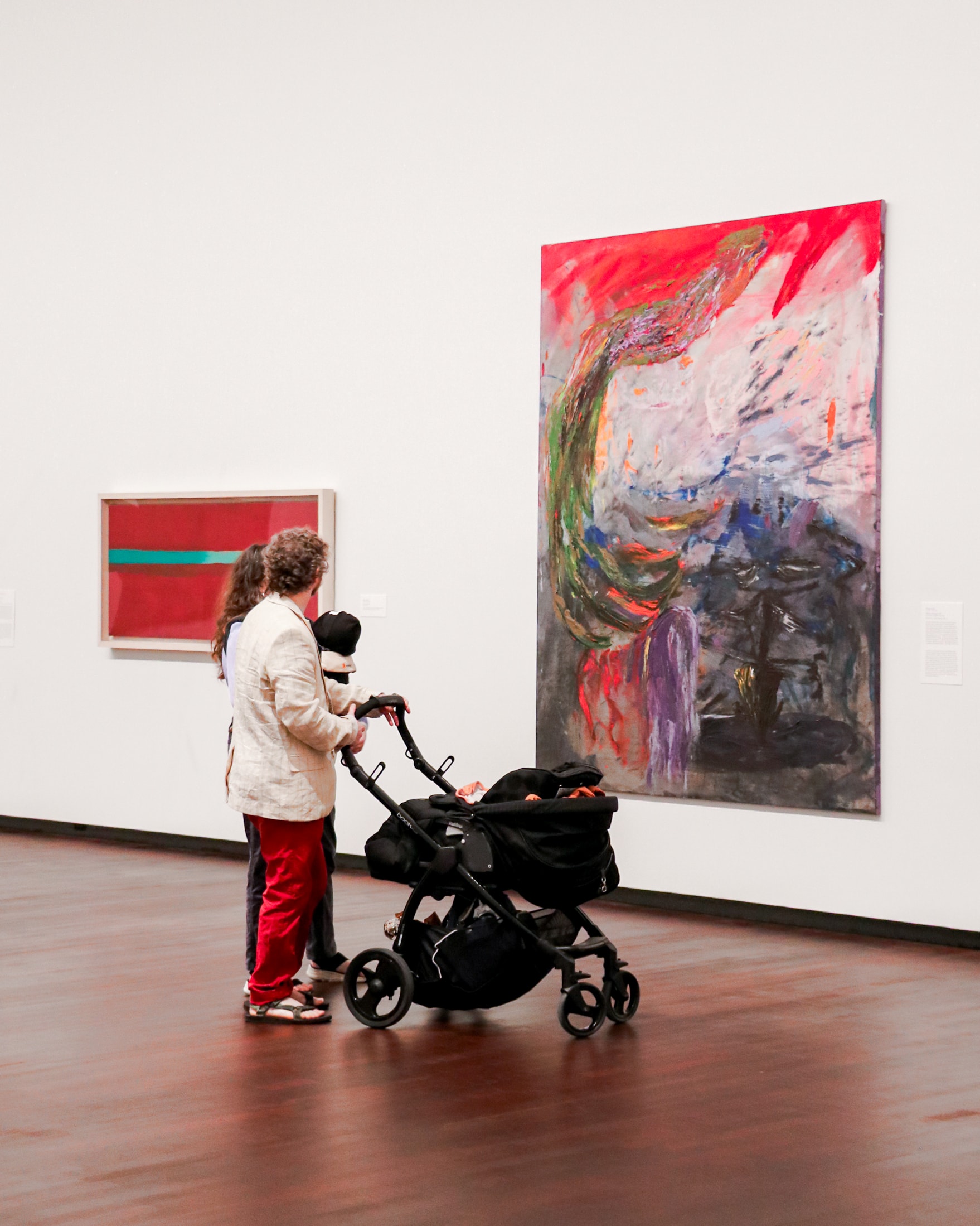 Two adults and a child in a stroller view colorful abstract paintings on the white walls of an art gallery with a polished wooden floor.