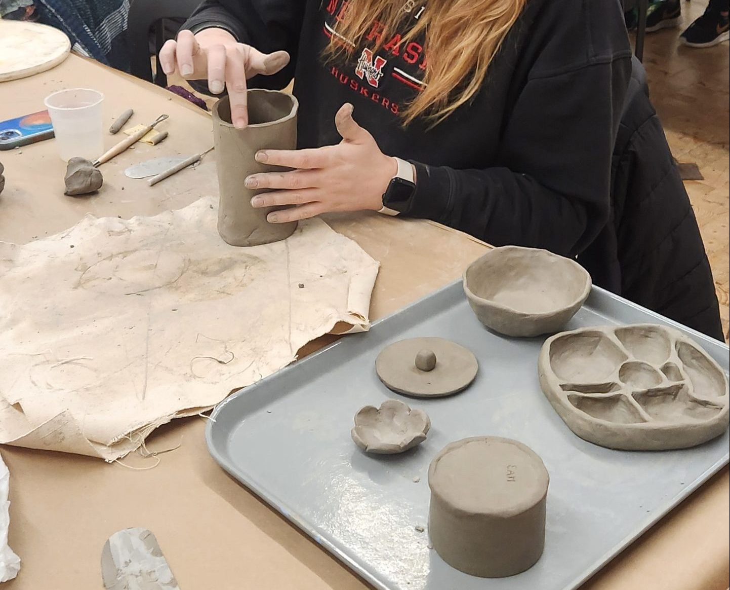 A woman is seated at a table engaged in pottery. She is shaping a clay cylinder. Various clay objects and tools are on a tray in front of her. Other people are in the background, also working on pottery.