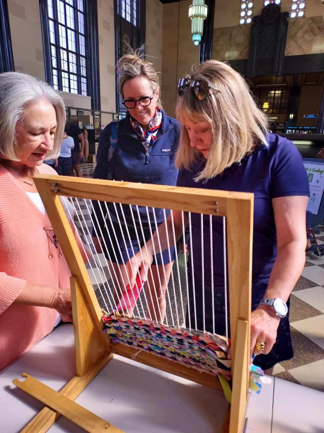 Three women work together on a wooden loom, weaving colorful fabric strips. They are in a large room with art deco elements, including tall windows and hanging lights. The atmosphere is collaborative and focused.
