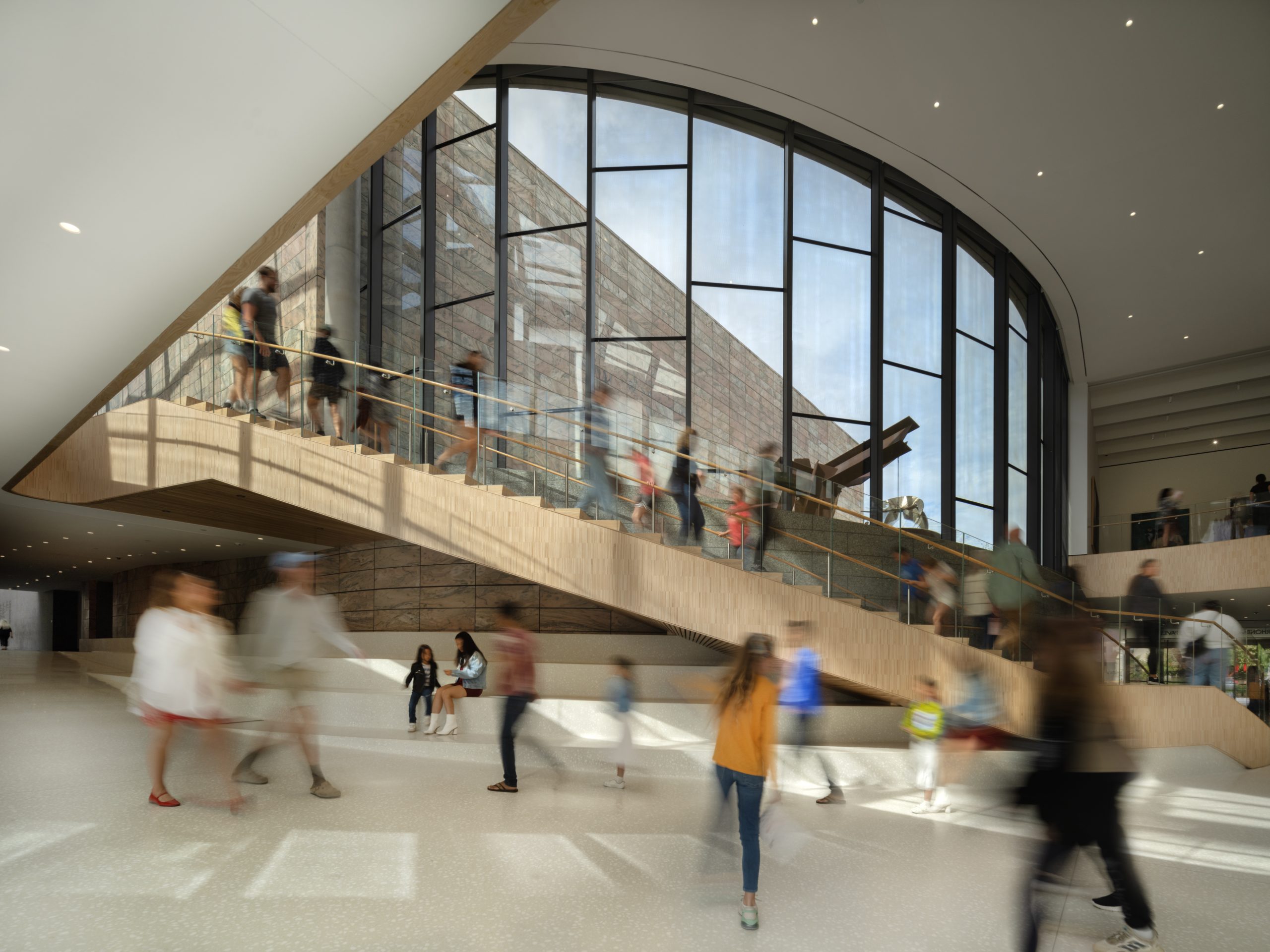 Spacious, modern interior with large arched windows and a wide staircase. Blurred figures move throughout, suggesting bustling activity. Natural light illuminates the space, highlighting the wooden accents and sleek design.