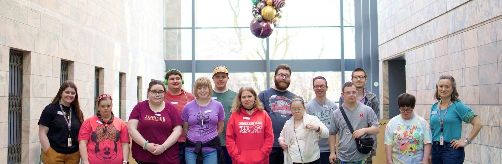 A group of thirteen people stand together indoors, smiling for a photo in front of large windows and a colorful sculpture hanging from the ceiling. Most are wearing casual clothes, and they appear to be in a bright, modern building.