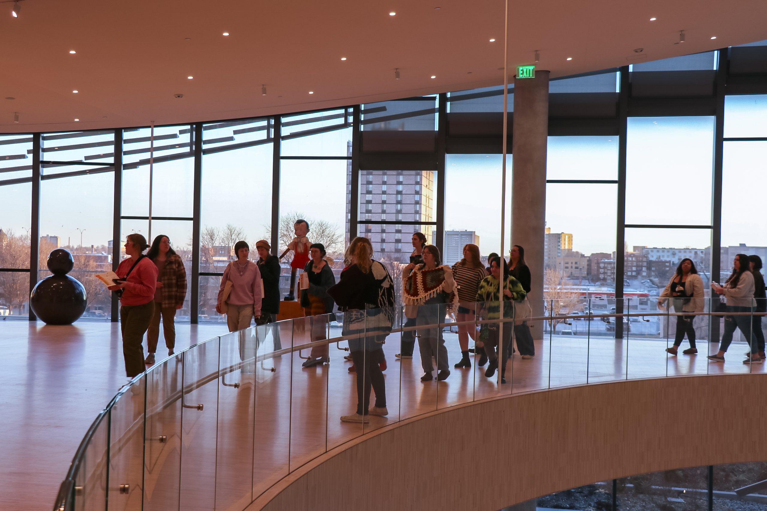 A group of people stand and walk along a curved glass railing inside a modern building with large windows. The skyline of a city is visible through the windows, and a dark abstract sculpture is in the background.