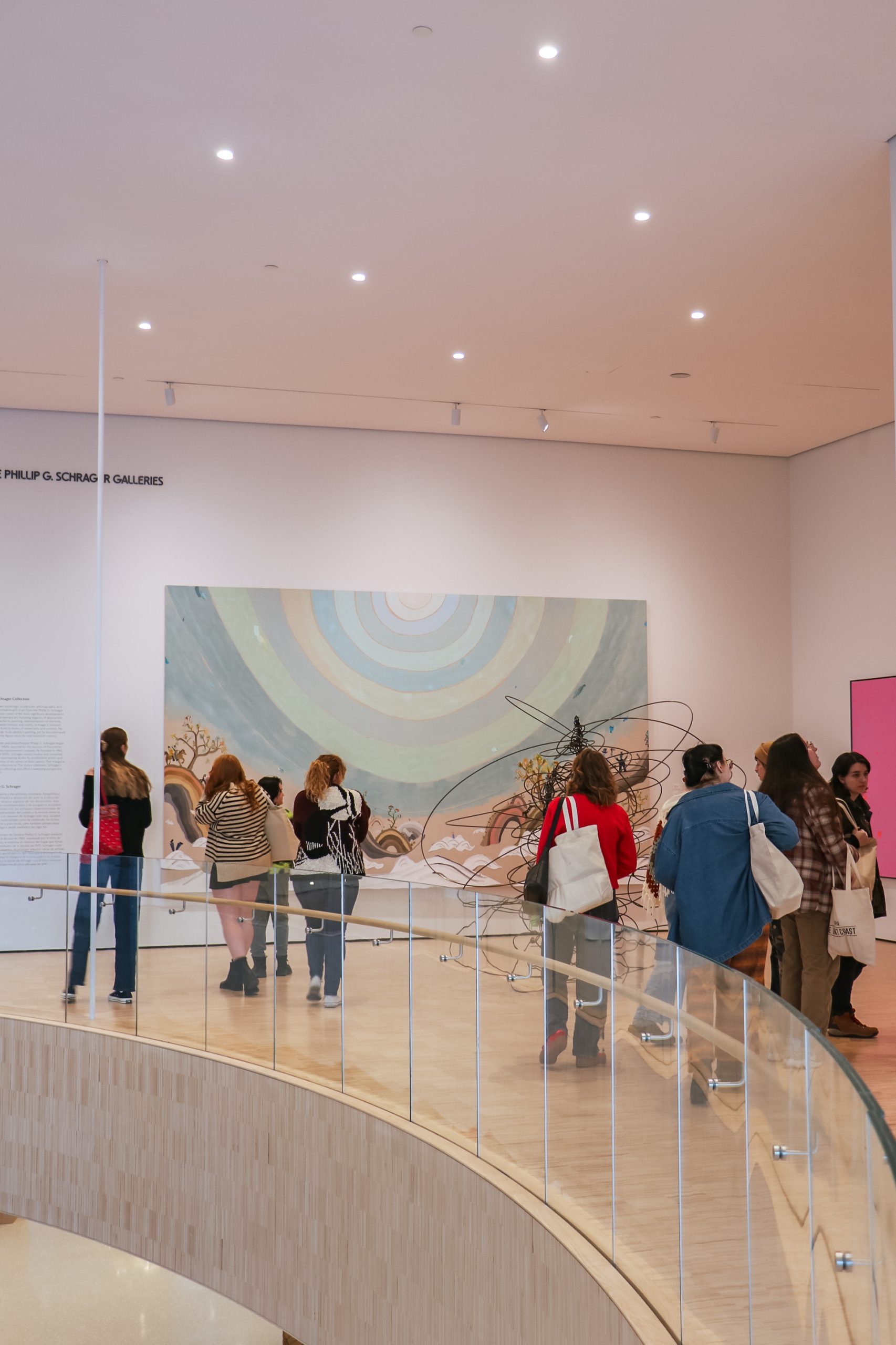 Visitors view artwork in a gallery with a large, abstract painting featuring circular patterns. The space has a curved walkway with a glass railing and bright lighting. Guests carry bags and wear casual clothing.