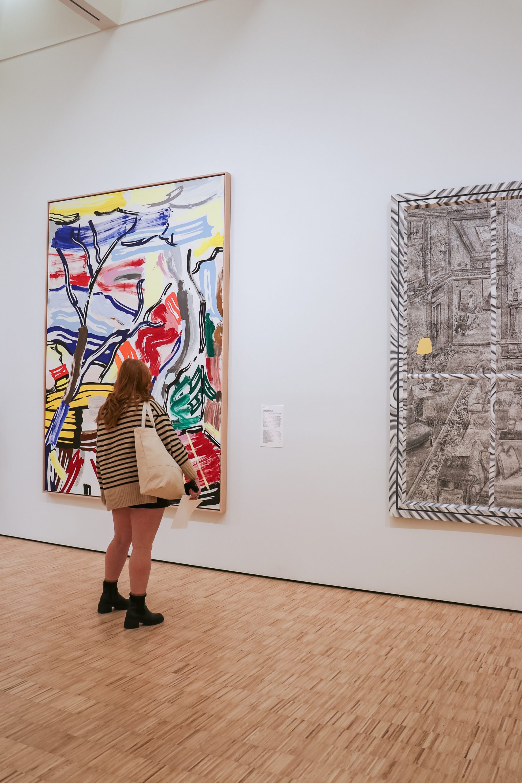 A person with long hair stands in an art gallery, looking at two large paintings. One is vibrant with abstract colors and shapes, while the other is more monochromatic with detailed line work. The gallery has wooden floors and white walls.