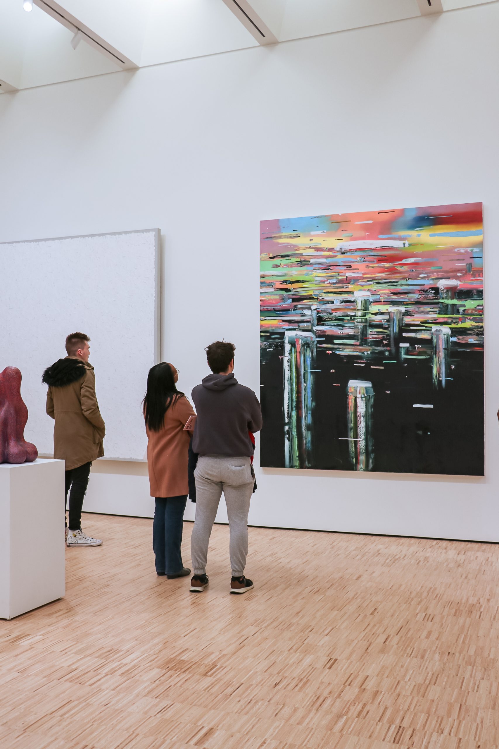 Three people stand in an art gallery, observing a large, colorful abstract painting on a white wall. The floor is wooden, and sculptures are displayed nearby. The painting features bright, horizontal streaks and reflective elements.