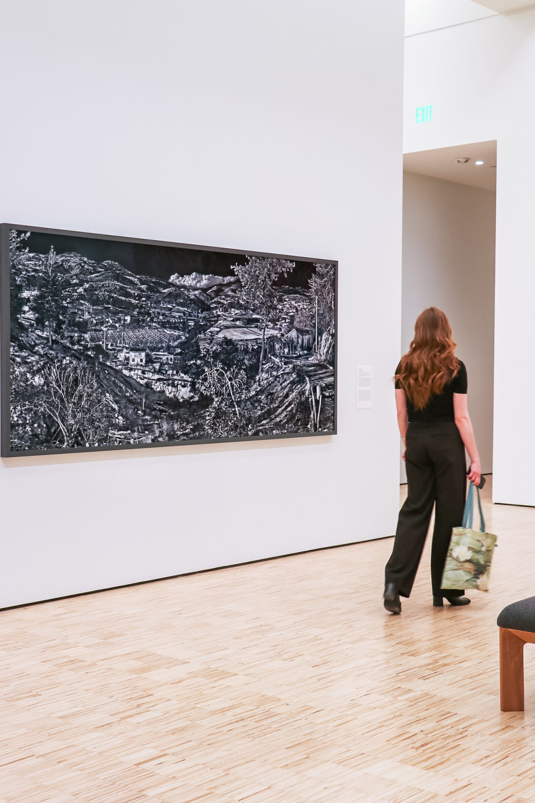 A person with long hair, wearing black clothing, walks through an art gallery. They are carrying a tote bag and are facing a large black and white landscape painting on the wall. The floor is wooden, and the setting is minimalist and spacious.
