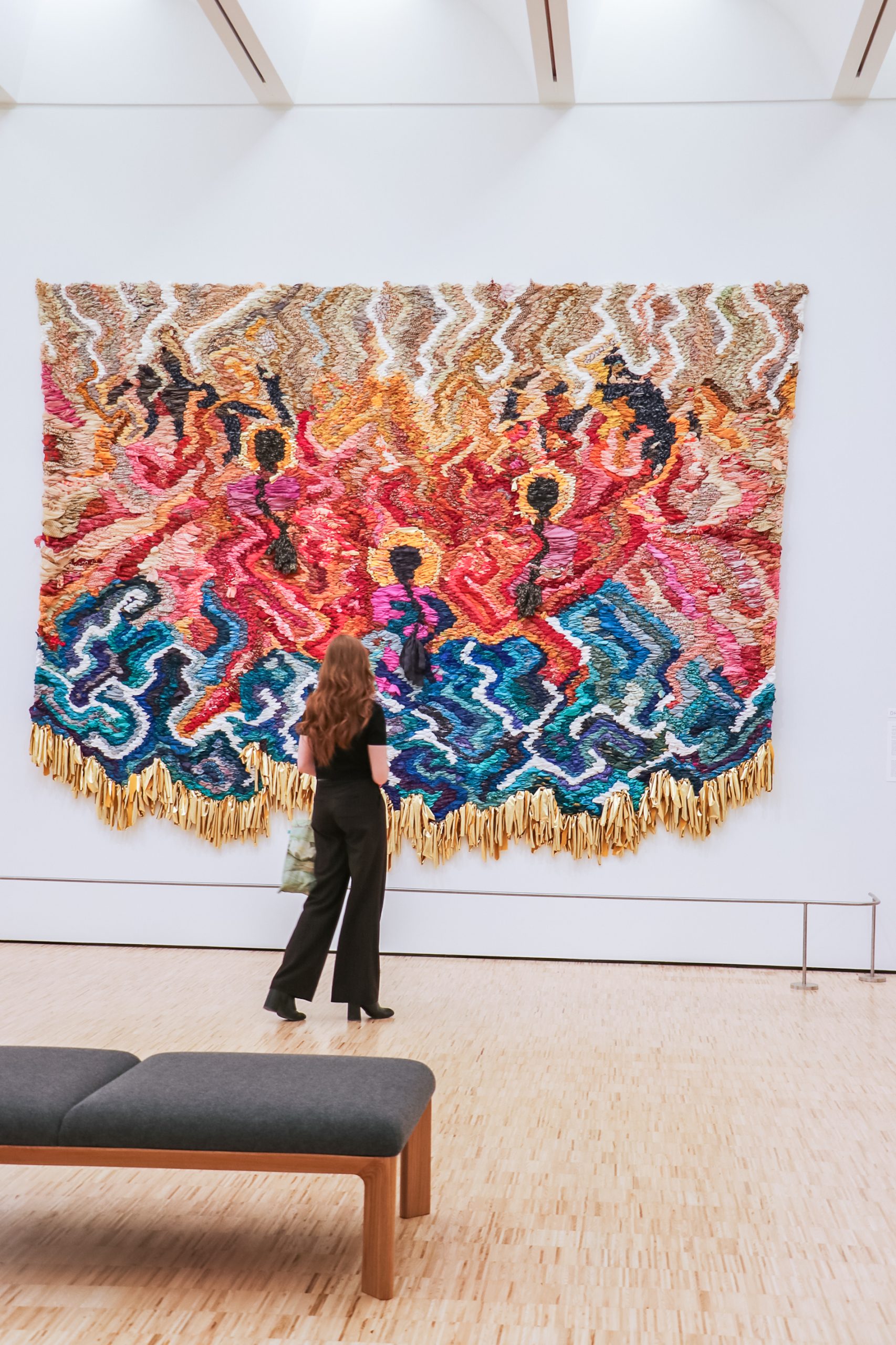 A person with long brown hair stands in front of a large, colorful woven tapestry featuring abstract patterns and figures. The artwork is displayed on a white gallery wall with a wooden floor and a bench in the foreground.