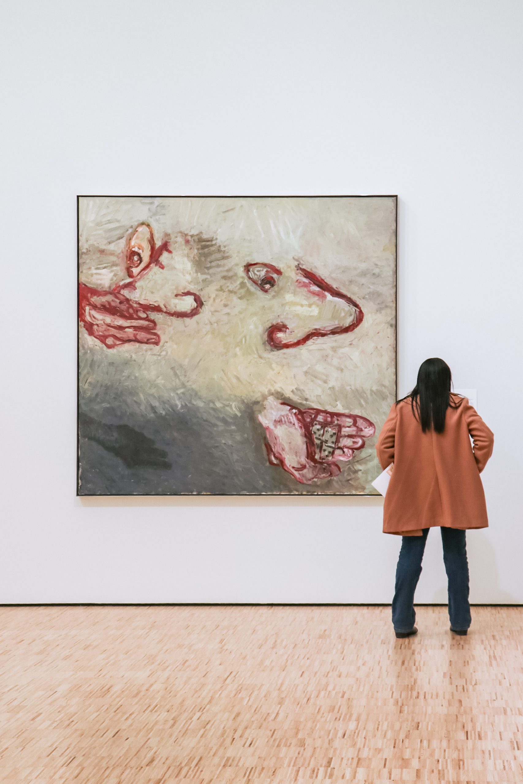 A person in a brown coat and jeans stands in front of a large abstract painting. The artwork features swirling colors and red, hand-like shapes on a textured background. The gallery floor is made of light wood.