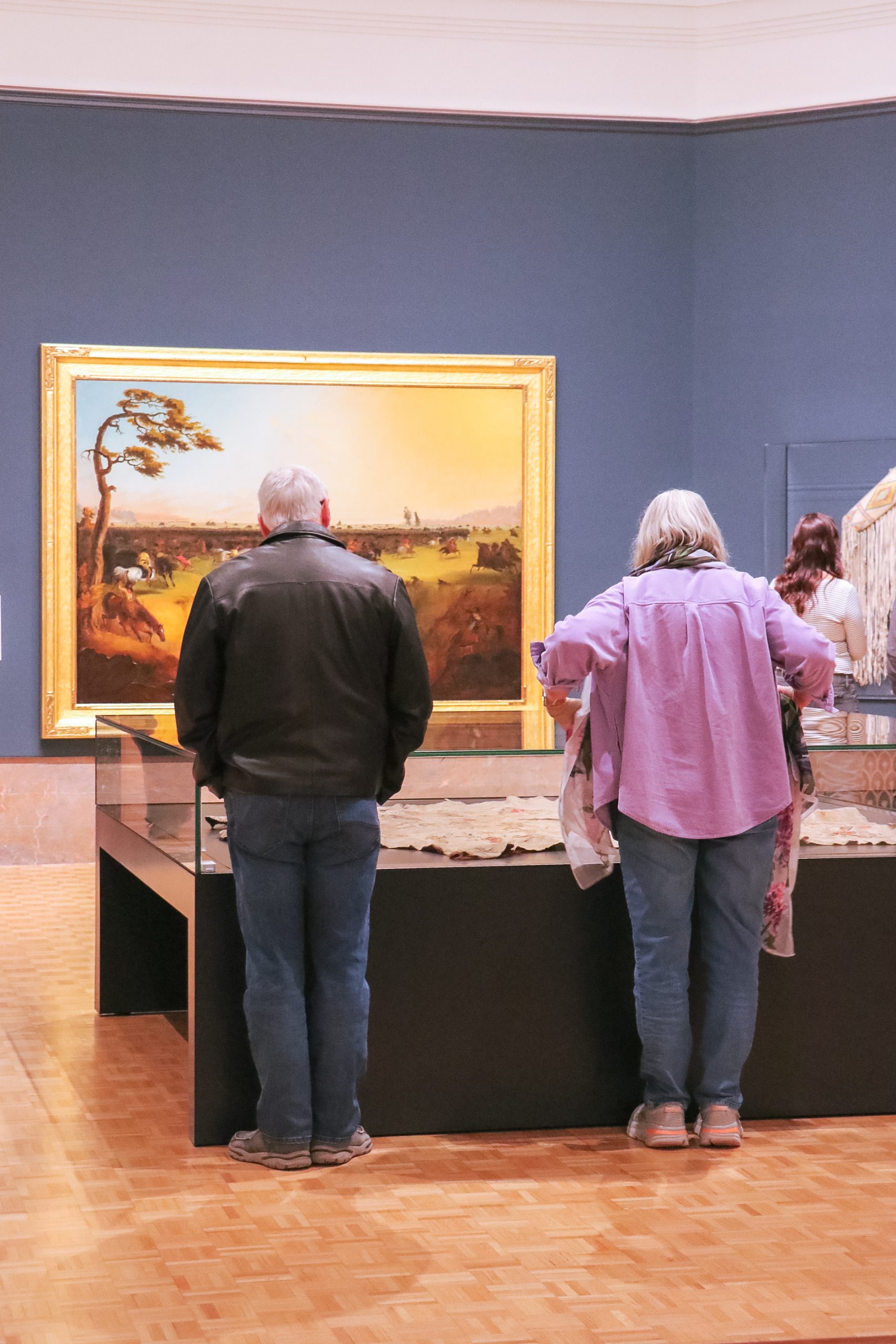 Two people stand closely viewing a large display case in an art gallery. A framed landscape painting hangs on the blue wall behind them. The room has a wooden floor and soft lighting.