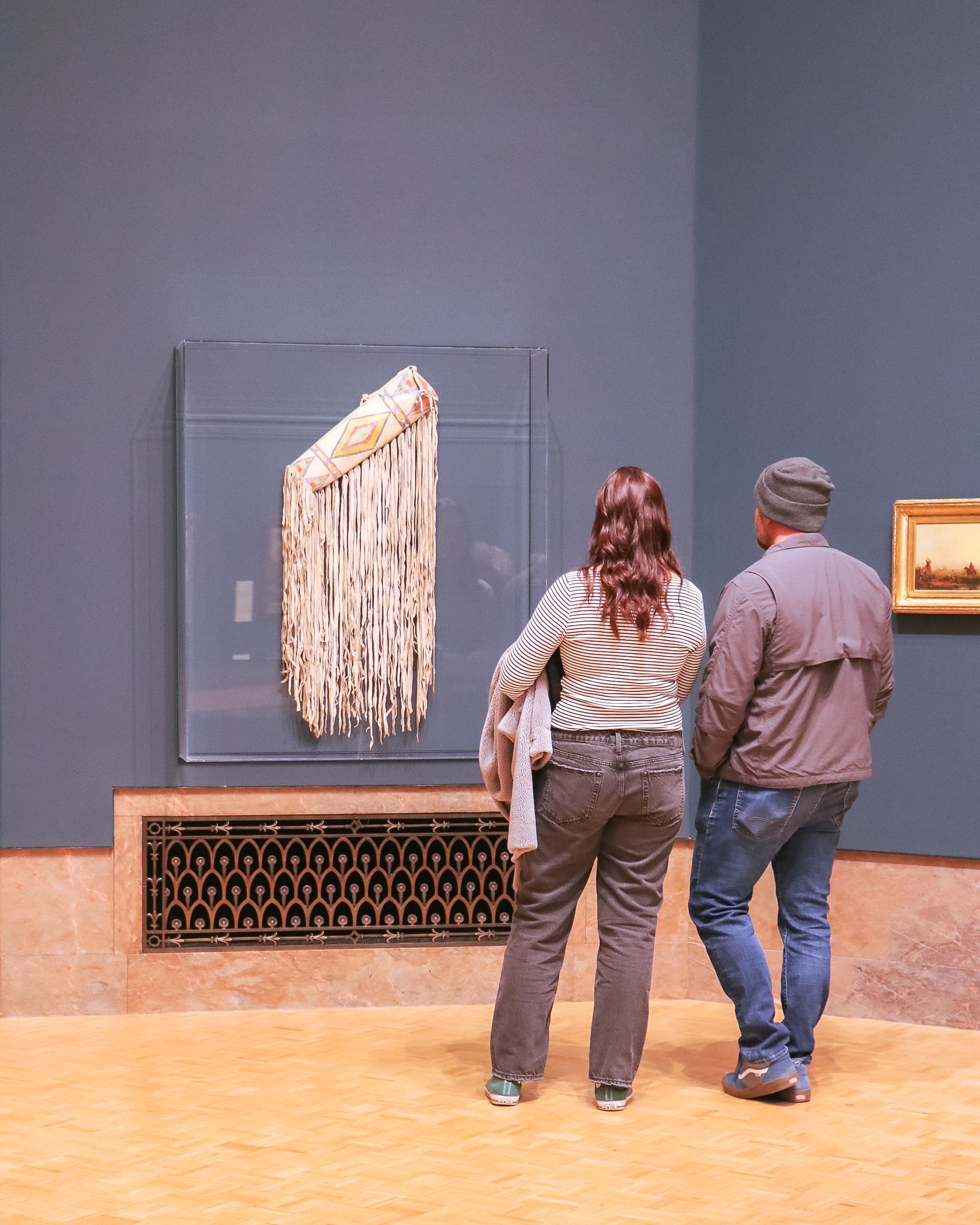 Two people stand in a museum, observing a fringed, beaded artifact displayed in a glass case. The room has blue walls and wooden flooring. A small framed painting hangs on the right. They are dressed casually.