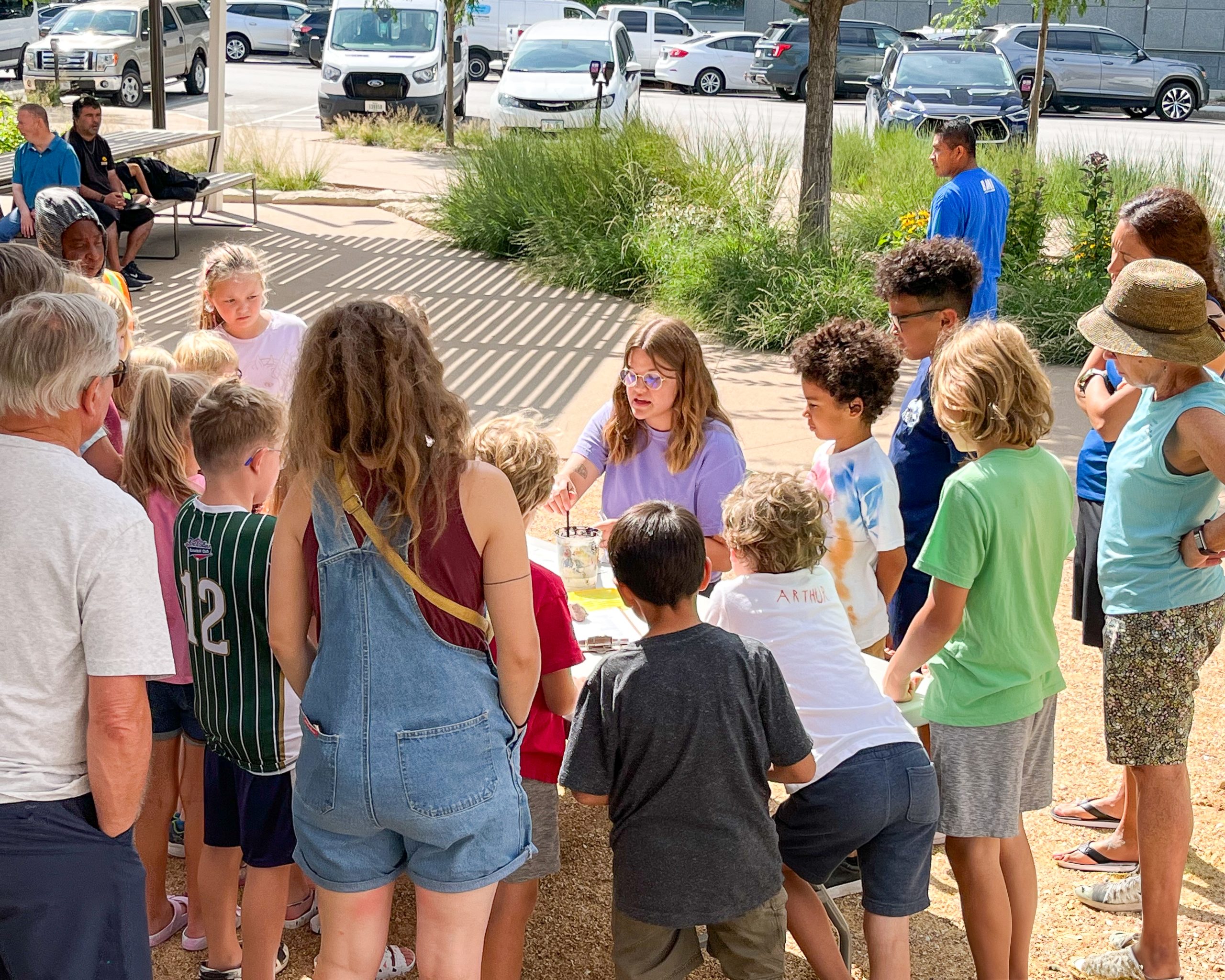 A group of children and adults gather around a table outside, watching a woman demonstrating something. The event takes place in a sunny area with parked cars and greenery in the background. The atmosphere appears educational and interactive.