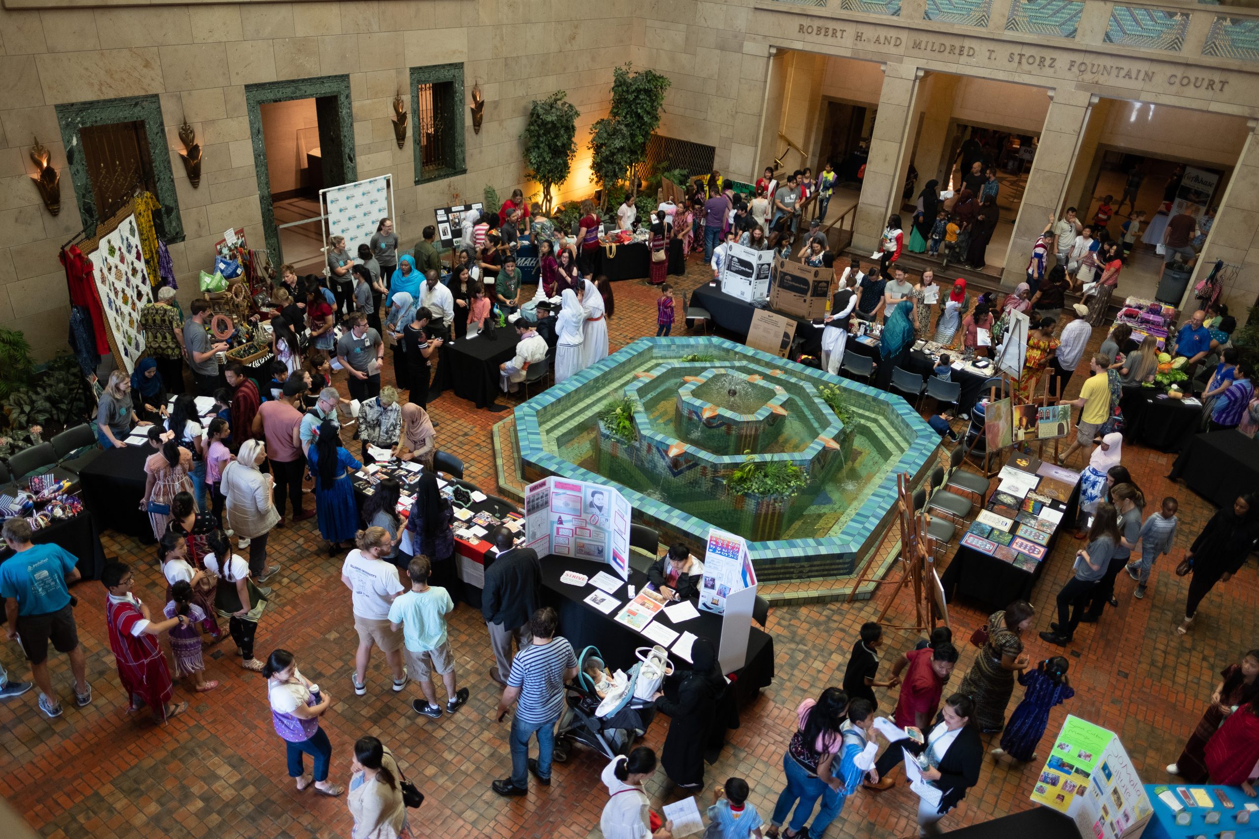 A large indoor event with many people visiting tables around a central decorative fountain, in a spacious hall with high ceilings, tiled floor, and vendors displaying various items and posters.