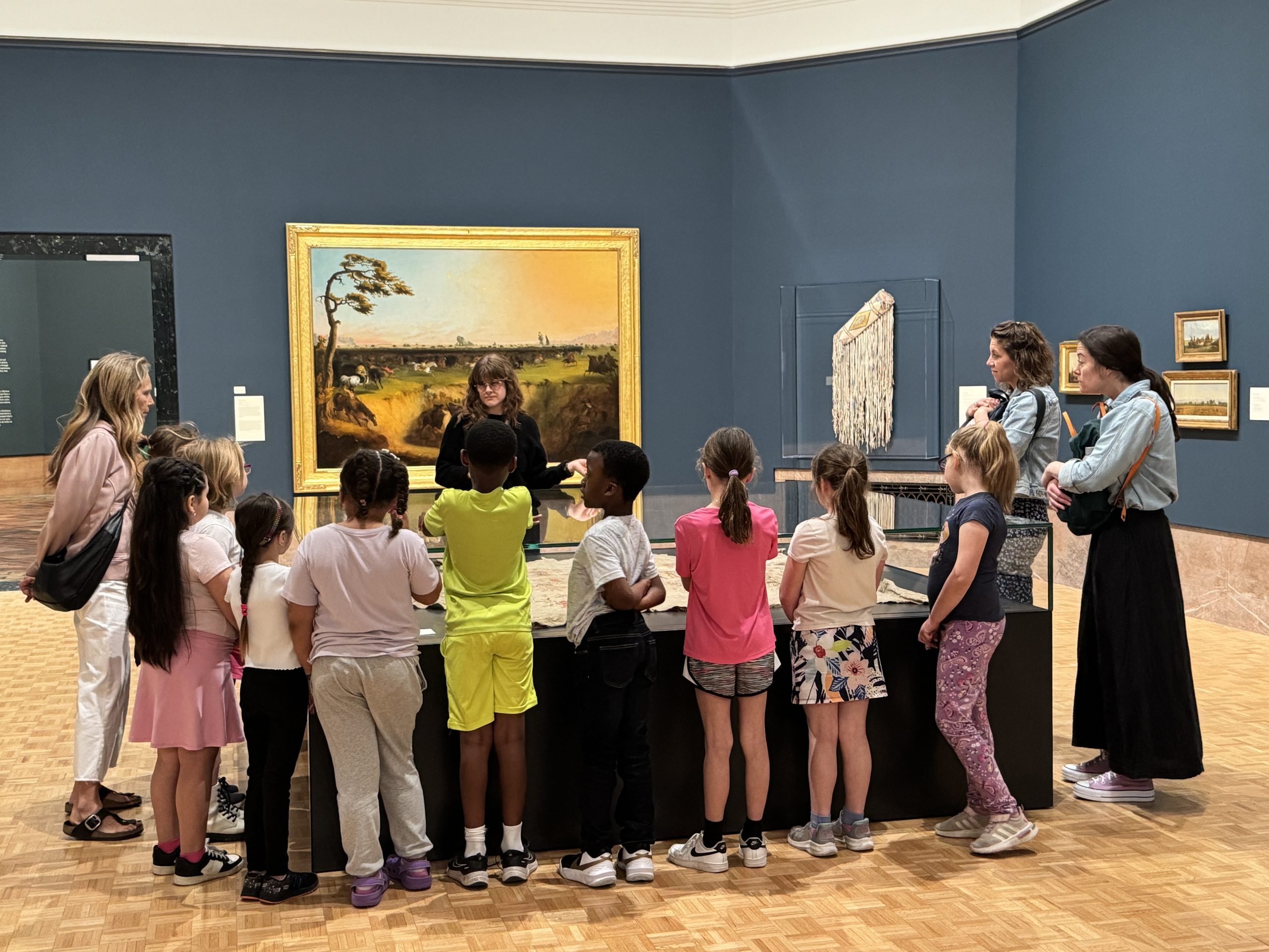A group of children and adults listen to a guide in an art museum. They stand around a display table, with paintings and sculptures visible on the blue walls behind them.