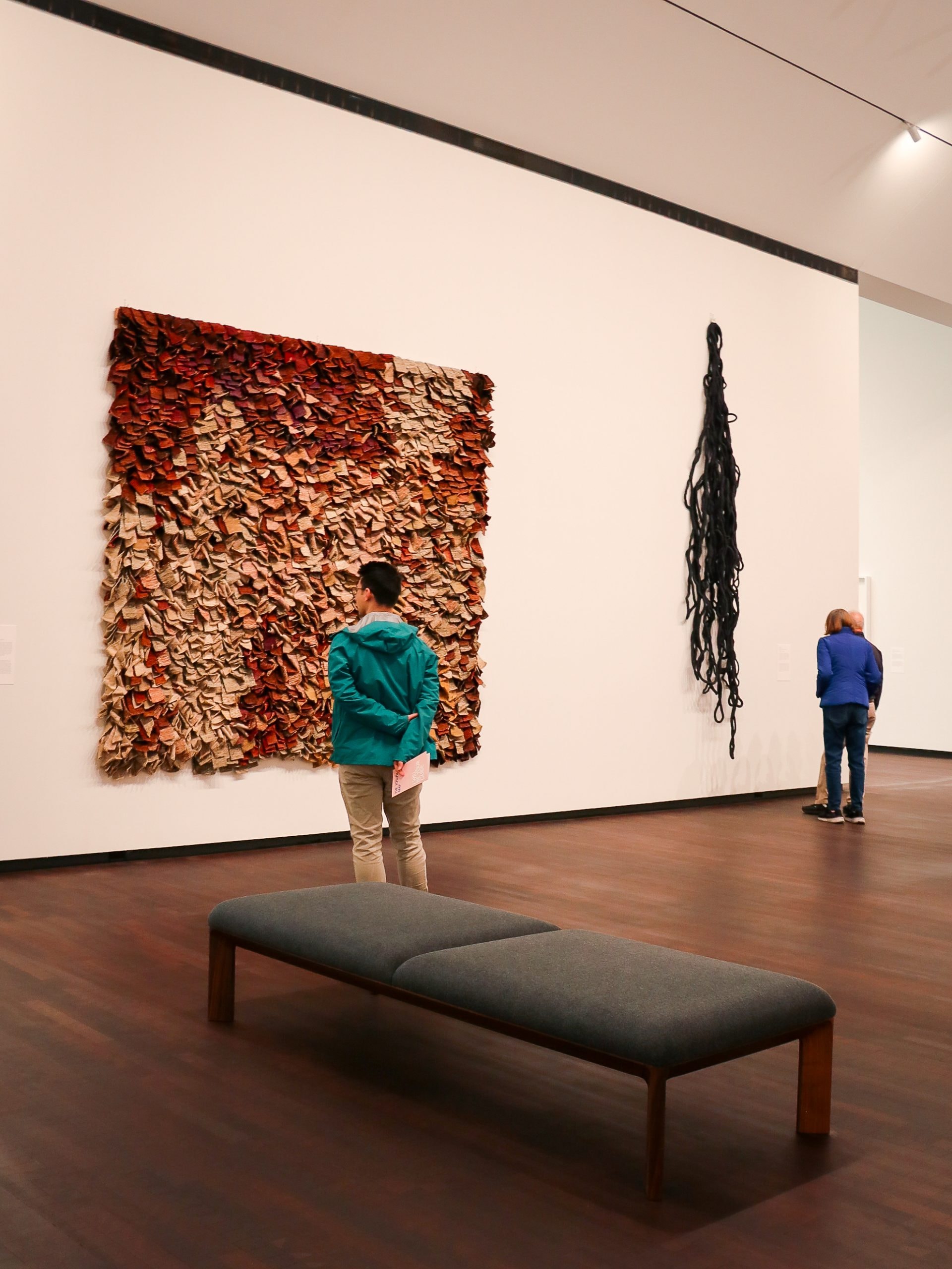 Two people view contemporary wall art in a gallery; one artwork is a textured, layered piece in browns and reds, and the other is a black, hanging installation. A bench sits in the foreground.