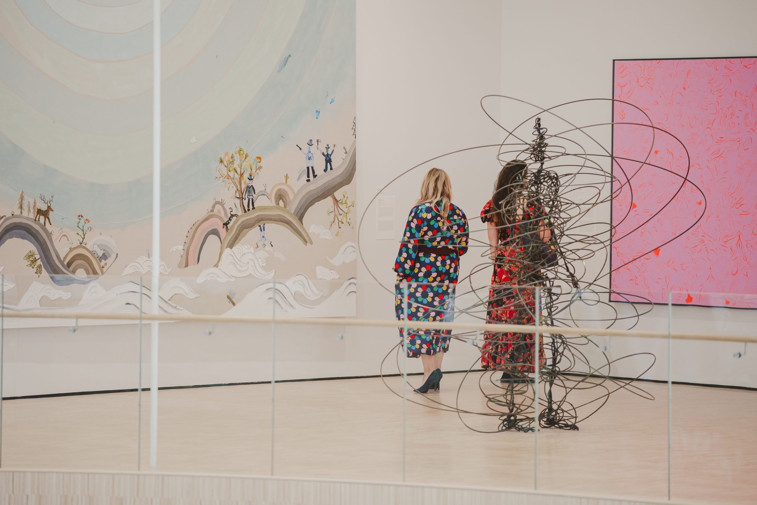 Two women in colorful dresses walk through an art gallery with abstract paintings on the walls and a modern wire sculpture in the foreground. The space is bright and open with light flooring and a glass railing.