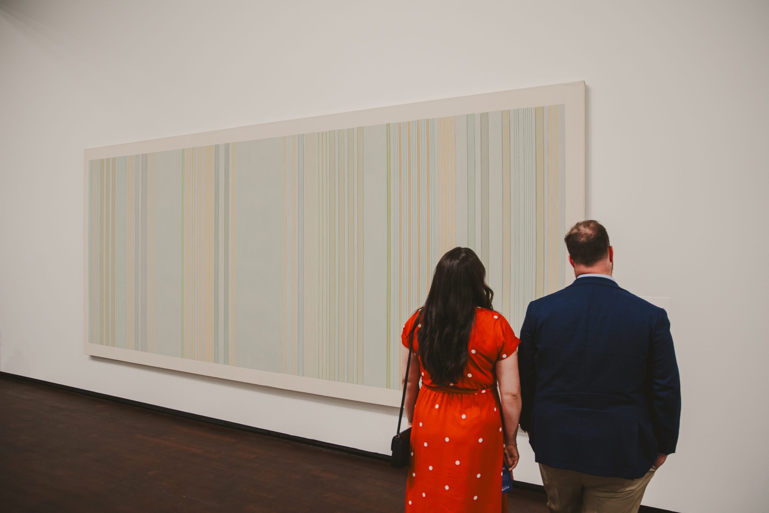 A woman in a red dress and a man in a blue jacket stand side by side in front of a large, striped abstract painting displayed on a white wall in an art gallery.