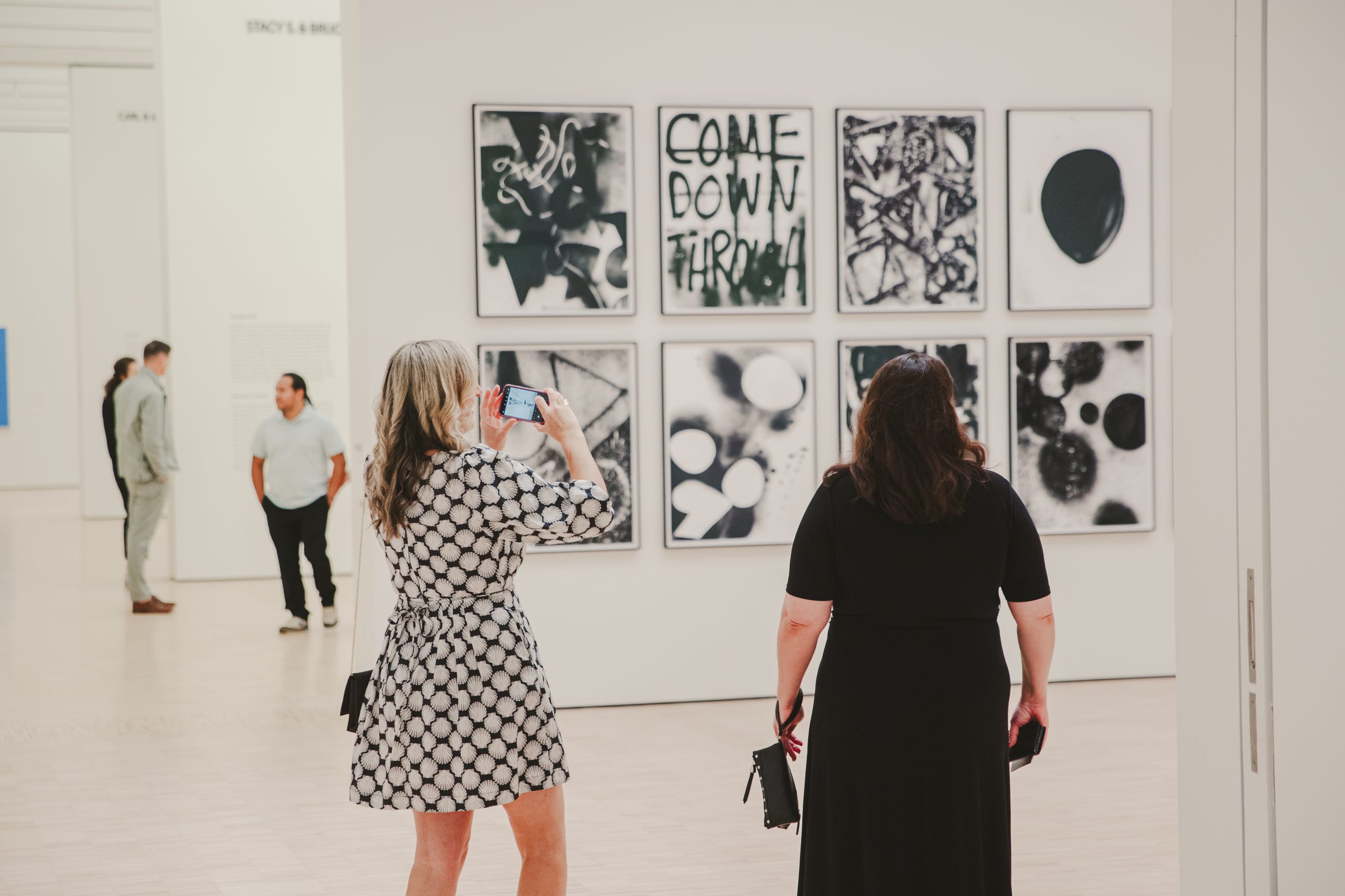 Two women stand in an art gallery; one is taking a photo of a wall displaying nine abstract black-and-white artworks, including one with the words "COME DOWN FROM." Other people are visible in the background.