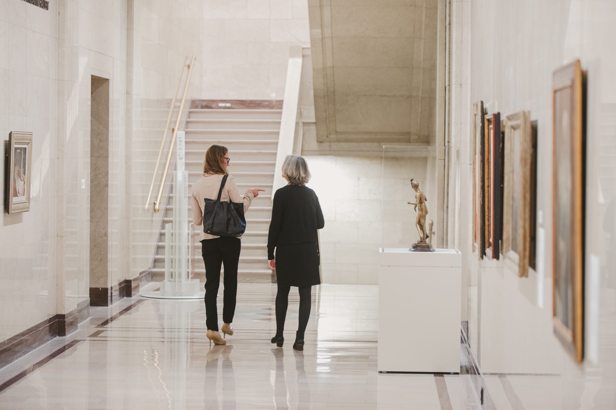 Two women walk down a bright, marble hallway in an art museum, engaged in conversation. Paintings hang on the walls and a small sculpture is displayed on a pedestal to their right.