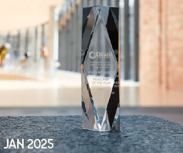 A clear, faceted glass award with engraved text stands on a textured surface. The engraving includes "OMAHA," "HEADLINER OF THE YEAR," and the year 2025. "JAN 2025" is written in bold text at the bottom left.