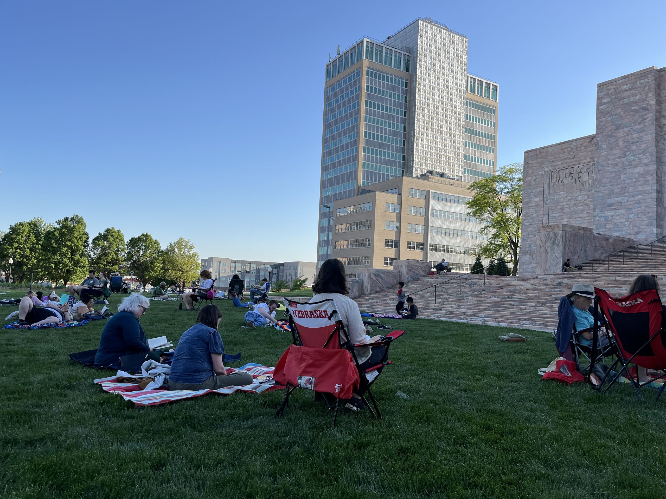 People sit on blankets and lawn chairs on a grassy area, enjoying a sunny day near a large stone monument and tall modern office buildings in the background. Some are reading or relaxing under a clear blue sky.