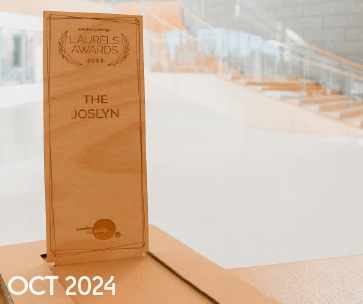 A wooden award plaque titled "THE JOSLYN" with "LAURELS AWARDS 2024" engraved, placed on a table. The background shows a modern staircase. "OCT 2024" is written in white in the bottom corner.