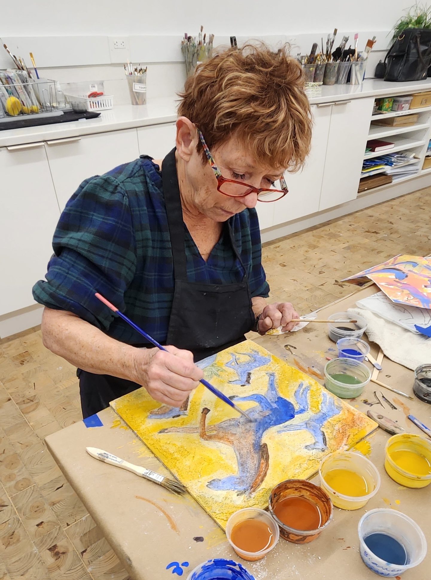 An older woman wearing glasses and a dark apron paints a colorful abstract design on a canvas at a table, surrounded by jars of paint in shades of blue, brown, yellow, and green in an art studio.