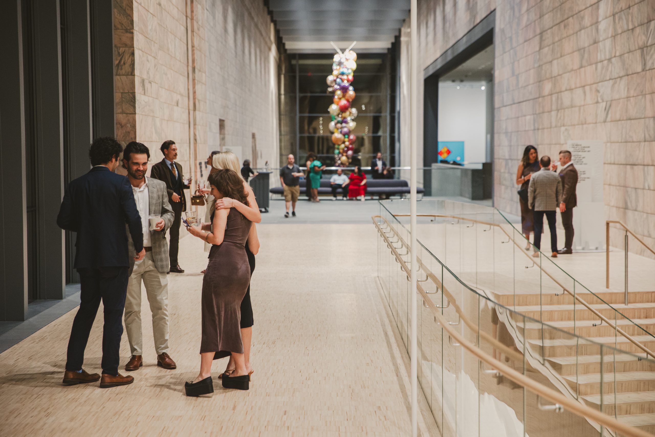 People dressed in semi-formal attire converse and mingle in a spacious modern art museum corridor with stone walls, glass railings, and colorful balloon-like sculpture at the far end.